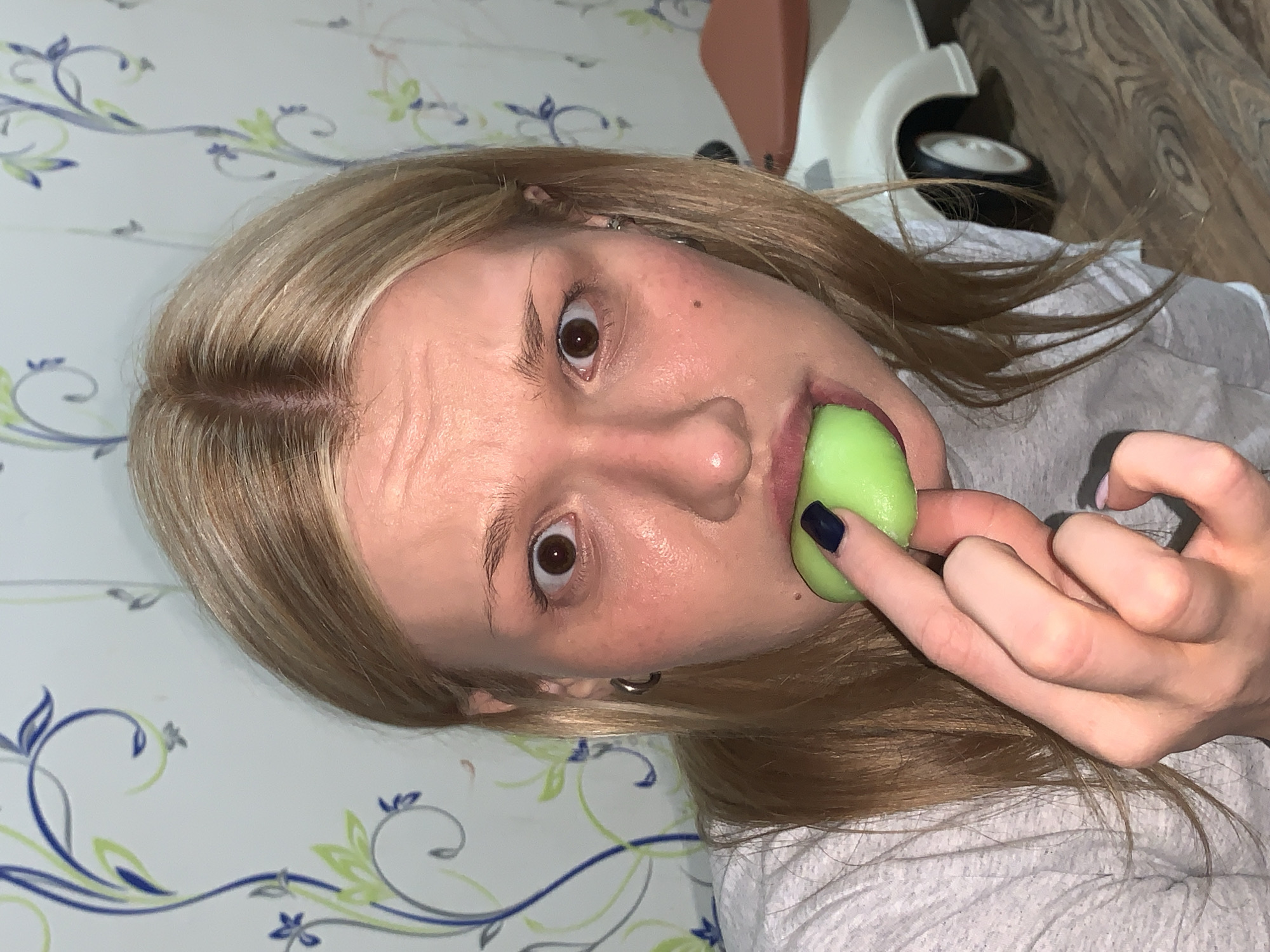 A light-skinned female with blonde hair is holding a green object in her mouth with one hand. She appears to be indoors. The background features a wallpaper with a floral design and wooden flooring. Behind the woman is a white object with a wheel that appears to be part of a child's toy. The location is unknown, and there is no visible evidence suggesting the location is Chaikovsky, Russia.