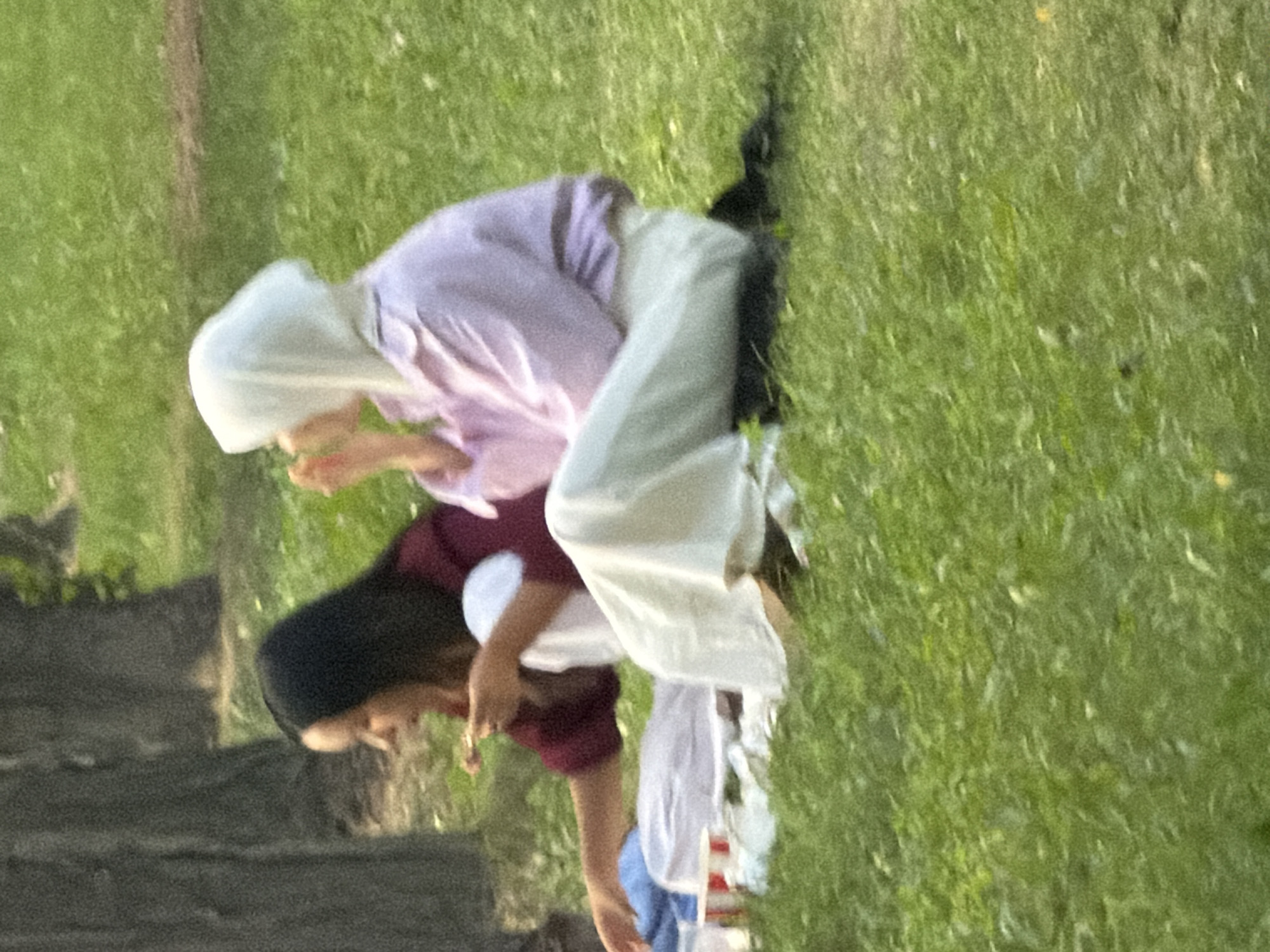 Two individuals are seated on a grassy lawn in front of a tree. The person on the left has dark hair and is wearing a maroon shirt and white pants. They are holding a piece of food in their right hand. The person on the right is wearing a white hijab and a light purple shirt with white pants. They appear to be bringing food to their mouth with their right hand. A picnic blanket and some containers are visible in front of the individuals. The scene is outdoors, with a natural, green environment.