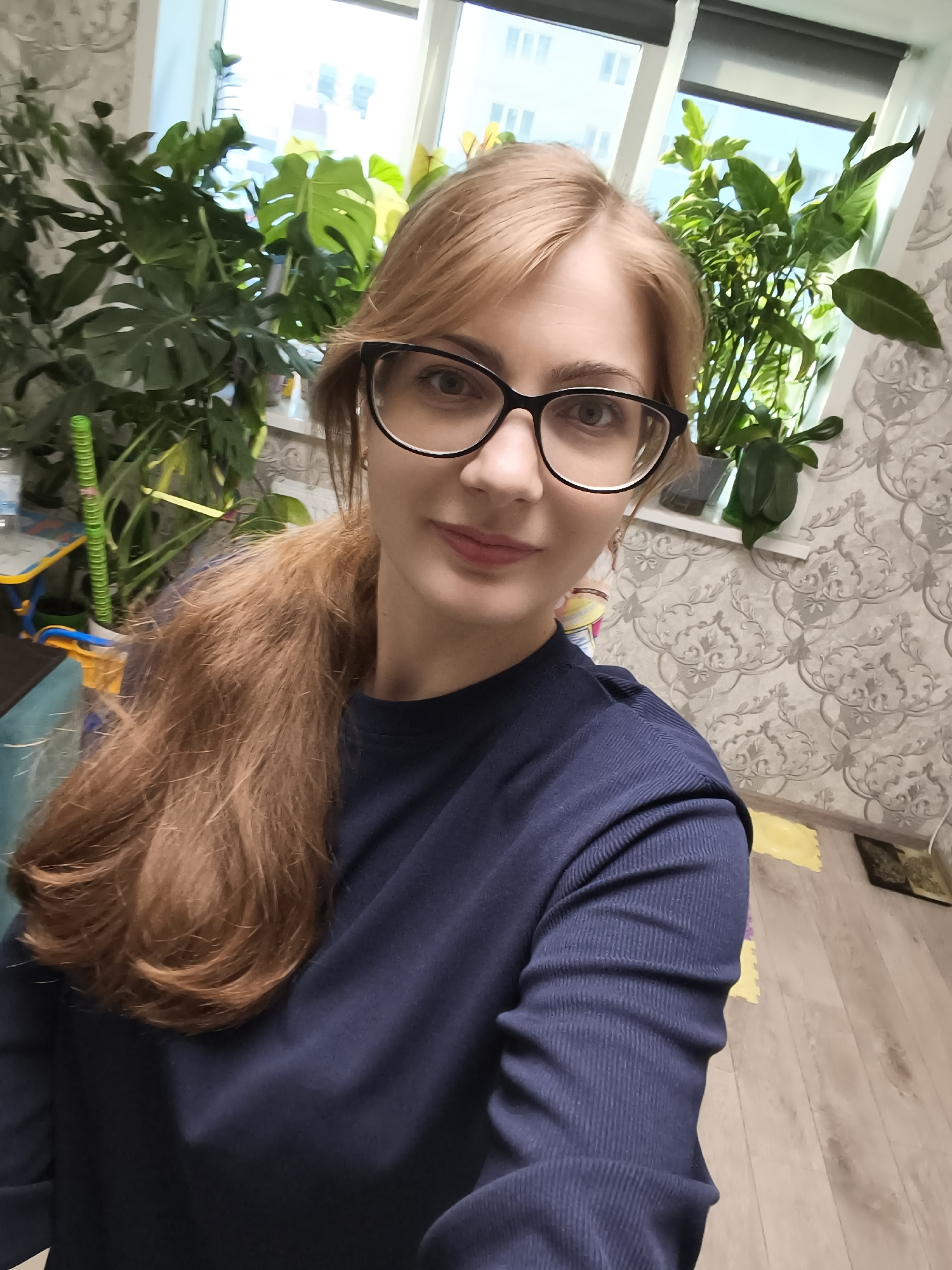 A woman with long, auburn hair tied in a ponytail and wearing black-rimmed glasses is taking a selfie. She has fair skin and is smiling slightly. She is wearing a dark blue, ribbed long-sleeved shirt. The background features several large green houseplants, a window with blinds, and patterned wallpaper. The flooring appears to be wood. The scene suggests an indoor setting, possibly a home. There is no visible text. The lighting and the presence of indoor plants suggest it is daytime. The location is identified as Novaya Usman', Russia.