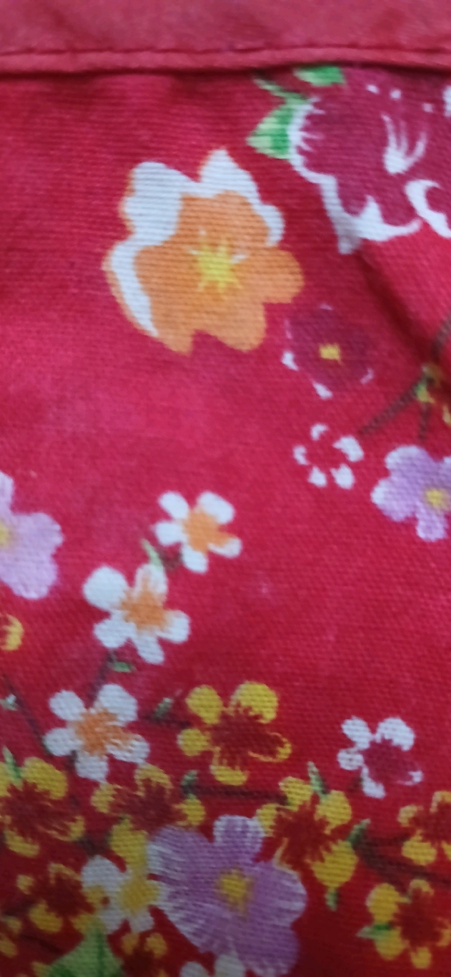 The image is a close-up, vertical, slightly blurry photograph of a red fabric with a floral print. The flowers are depicted in various colors, including white with orange centers, pink, yellow, and dark red. Some of the flowers have multiple petals, while others are smaller and star-shaped. Green leaves and thin brown stems are also visible. A dark red seam runs horizontally across the top of the image, suggesting this might be a hem or edge of a garment or textile. The focus is entirely on the pattern of the fabric, with no discernible background or environmental context. There are no people or specific objects identifiable beyond the fabric itself. The location in the city of Darussalam, Indonesia is not discernible from the visual information provided in the image.