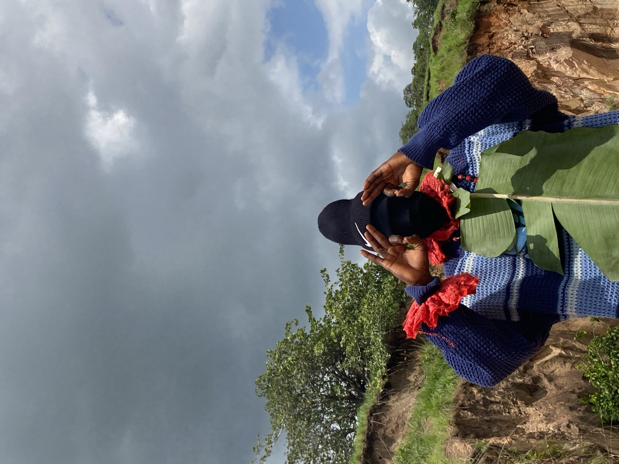 A person wearing a dark beanie, a black face mask, and a blue and white striped sweater stands on a grassy embankment. Red ruffled fabric adorns the person's neck, and large green leaves are attached to the front of their sweater. Their hands are raised, framing their face. Behind the person, a lush green tree grows on the embankment, which appears to be a ravine or cliff edge. The sky above is filled with dramatic, dark clouds, with patches of lighter sky and white clouds visible. The setting is outdoors, in a natural, possibly rural, environment. This scene is located in the city of Ufu Esioni, Nigeria.