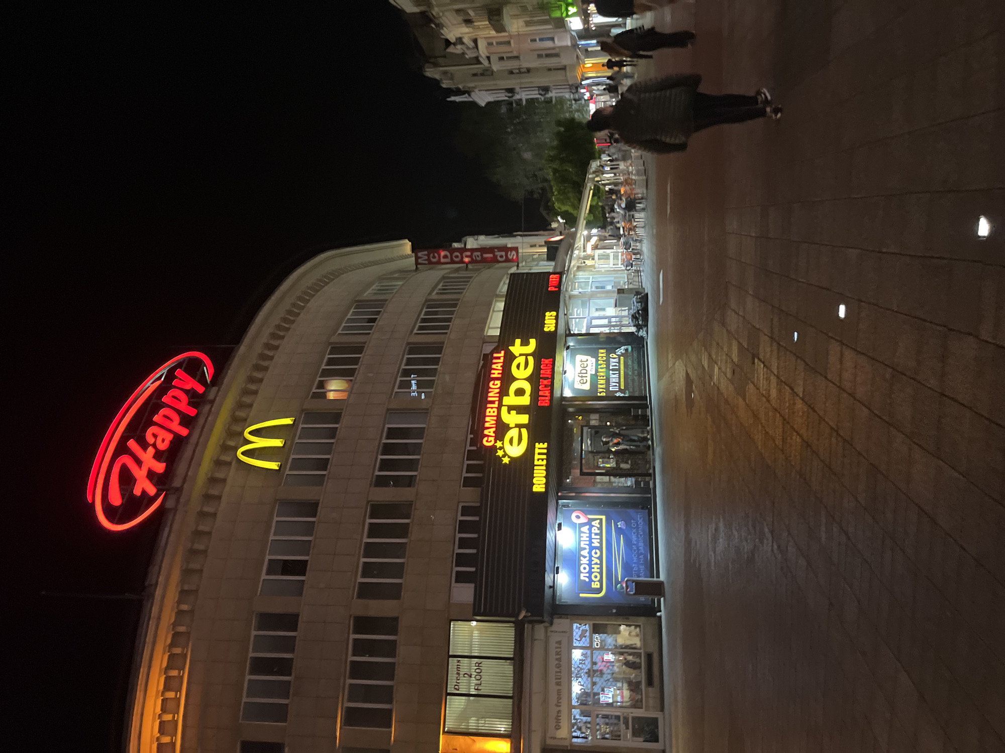 The image shows a street scene at night in Plovdiv, Bulgaria. A large curved building dominates the left side of the frame. The building features signs for Happy, McDonald's, and "efbet" (a gambling hall), and numerous windows. A pedestrian is walking along a wide tiled pavement in front of the building. Shops and other establishments line the street, indicated by illuminated signs and visible interiors. There are lights on the walkway. The sky is dark, suggesting it is nighttime.