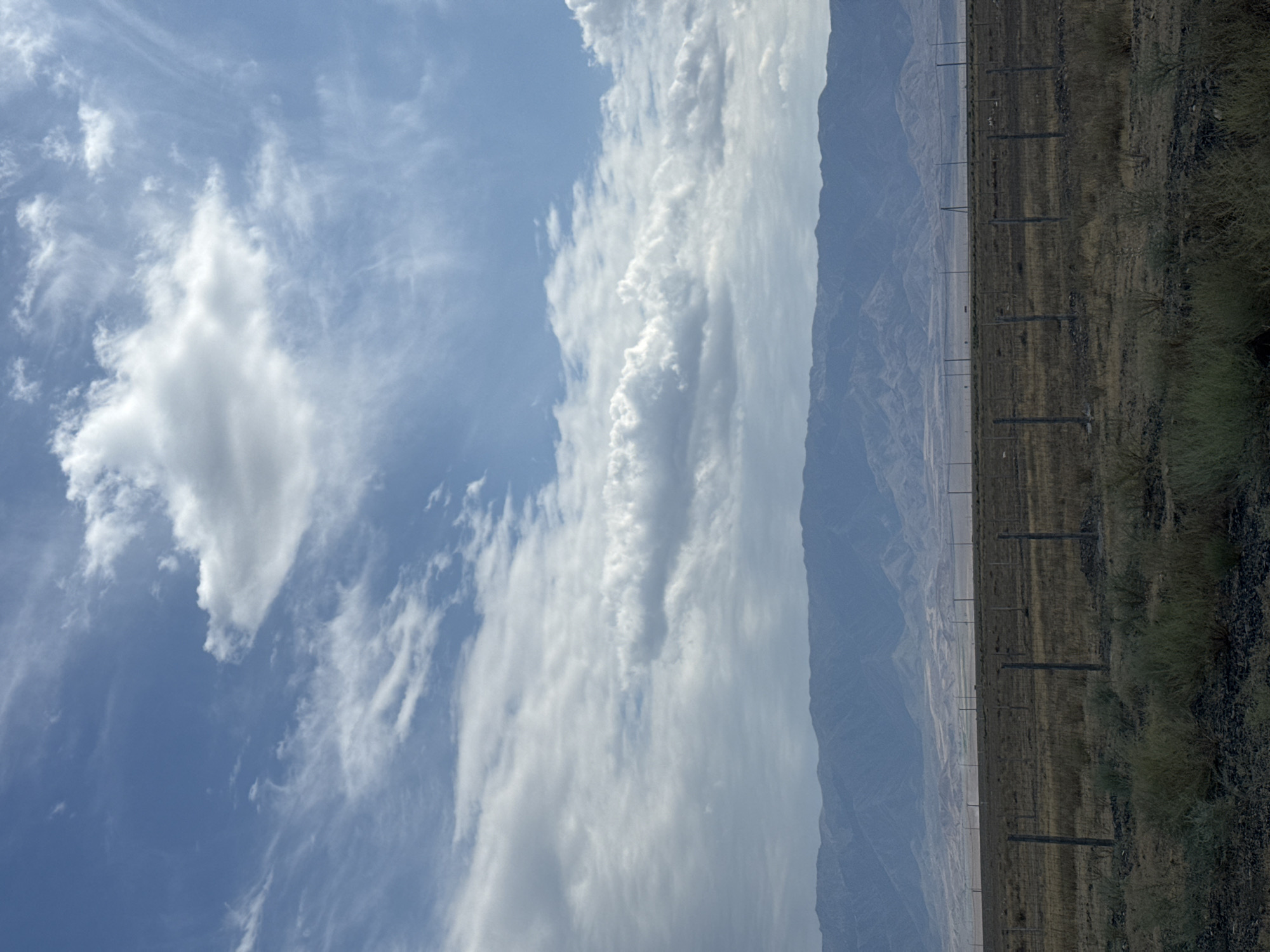 The image depicts a vast, arid landscape under a dynamic sky. In the foreground, a dry, grassy field is sectioned by a wire fence supported by wooden posts, suggesting a contained area or boundary. The field appears sparsely vegetated with dry grasses and some low-lying shrubs. Beyond the fence, the terrain stretches out to meet a range of distant, muted blue mountains that form the horizon. The sky above is a mix of deep blue and white, with scattered clouds ranging from wispy cirrus formations high up to larger, denser clouds closer to the mountain range. The overall impression is one of an open, natural environment with signs of human demarcation. The location is identified as Ақши, Kazakhstan.