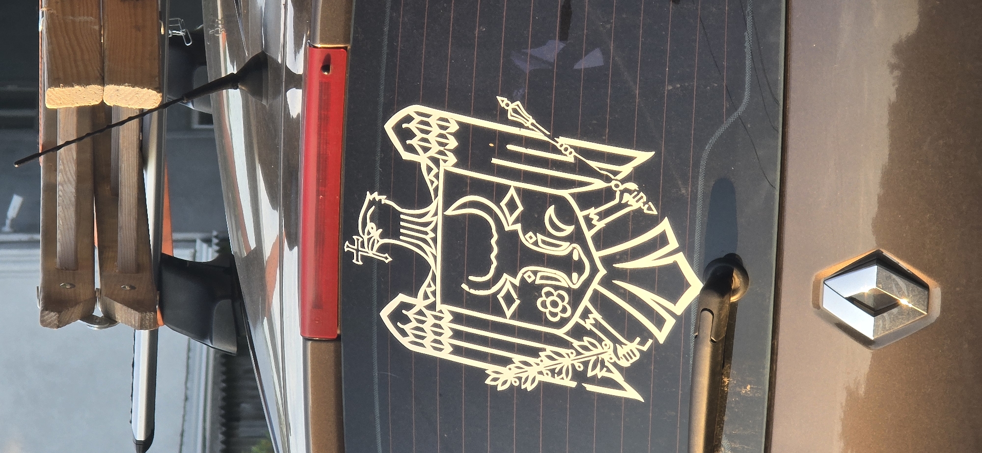 The image shows the rear of a dark-colored car, likely a hatchback, parked outdoors. A silver diamond-shaped emblem, the Renault logo, is visible on the trunk. A white decal of a coat of arms is affixed to the rear window, featuring an eagle with a cross above its head, a bull's head in the center, and elements like a flower and a scepter. A car antenna is visible on the roof, along with a wooden roof rack holding some items. The background suggests an urban or suburban setting with some buildings visible. Without further information, the specific location in Chișinău, Moldova, cannot be determined.