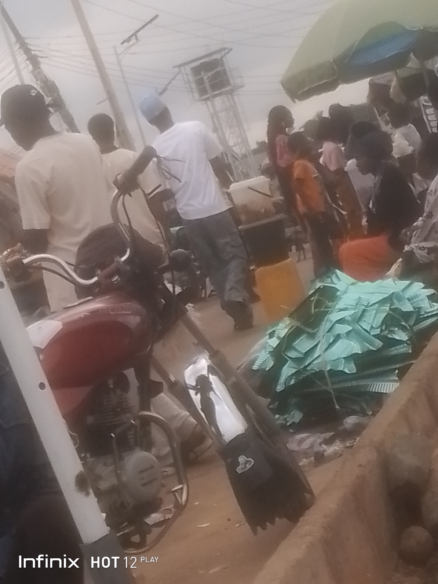 The image shows an outdoor scene, possibly a street market, with several people and a motorcycle. The lighting is overcast. In the foreground, a red motorcycle is partially visible, with its engine and handlebars clearly seen. To the right of the motorcycle, a large pile of green material, possibly roofing sheets or similar items, is covered with a blue tarp. Several people are seen in the background, some walking and some standing. One person is wearing a white t-shirt and a blue cap. Another person is wearing an orange shirt. The environment appears to be a dusty or dirt road. In the far background, there are utility poles and a structure that looks like a water tower. The Infinix HOT 12 PLAY logo is visible in the bottom left corner.