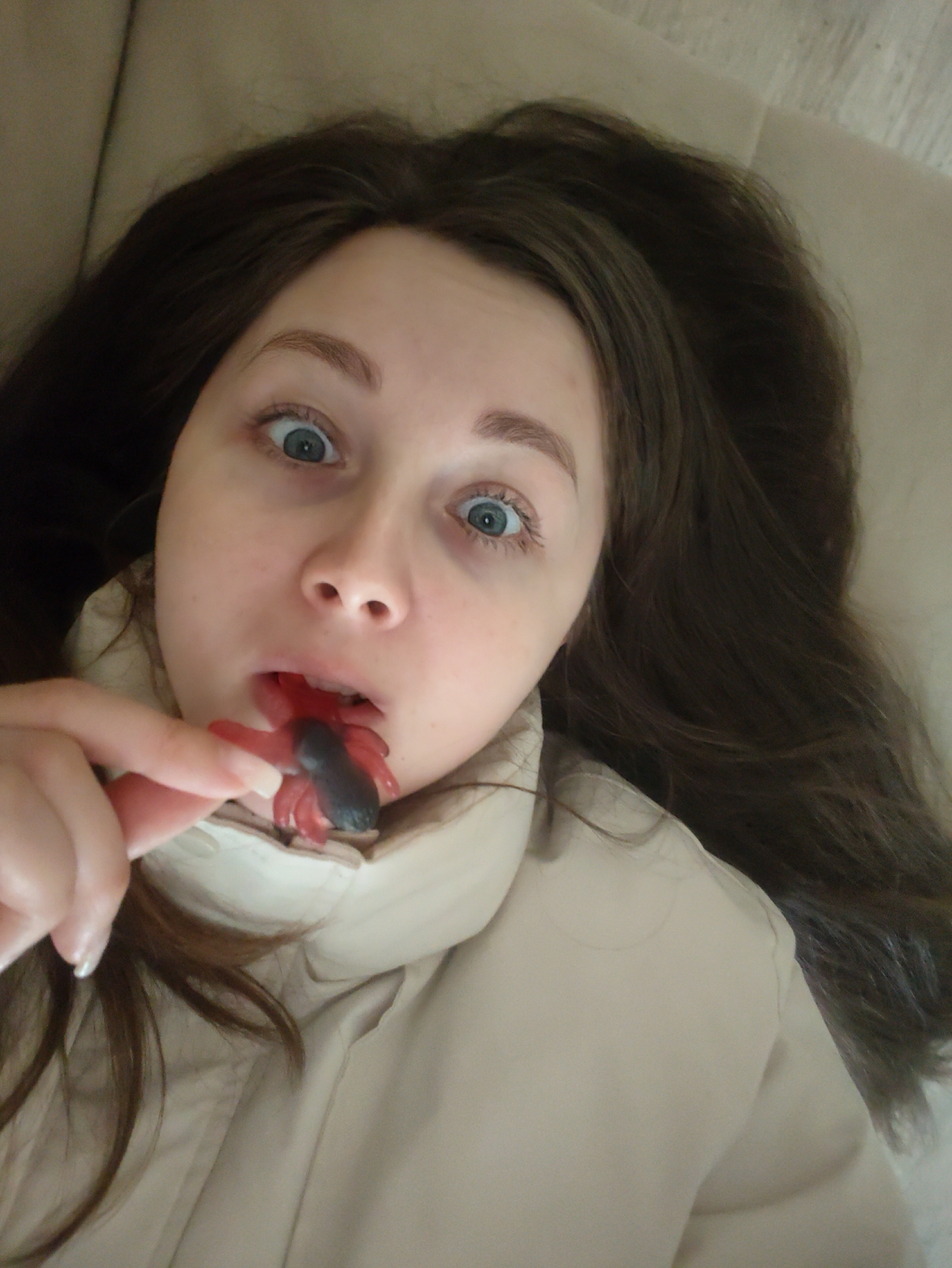 A fair-skinned young woman with brown hair and wide blue eyes is holding a red and black gummy candy resembling a bug towards her open mouth. She is wearing a beige, down-filled jacket. The background is blurry, suggesting she is indoors and possibly reclined on a soft surface. The setting appears to be an Unknown location.