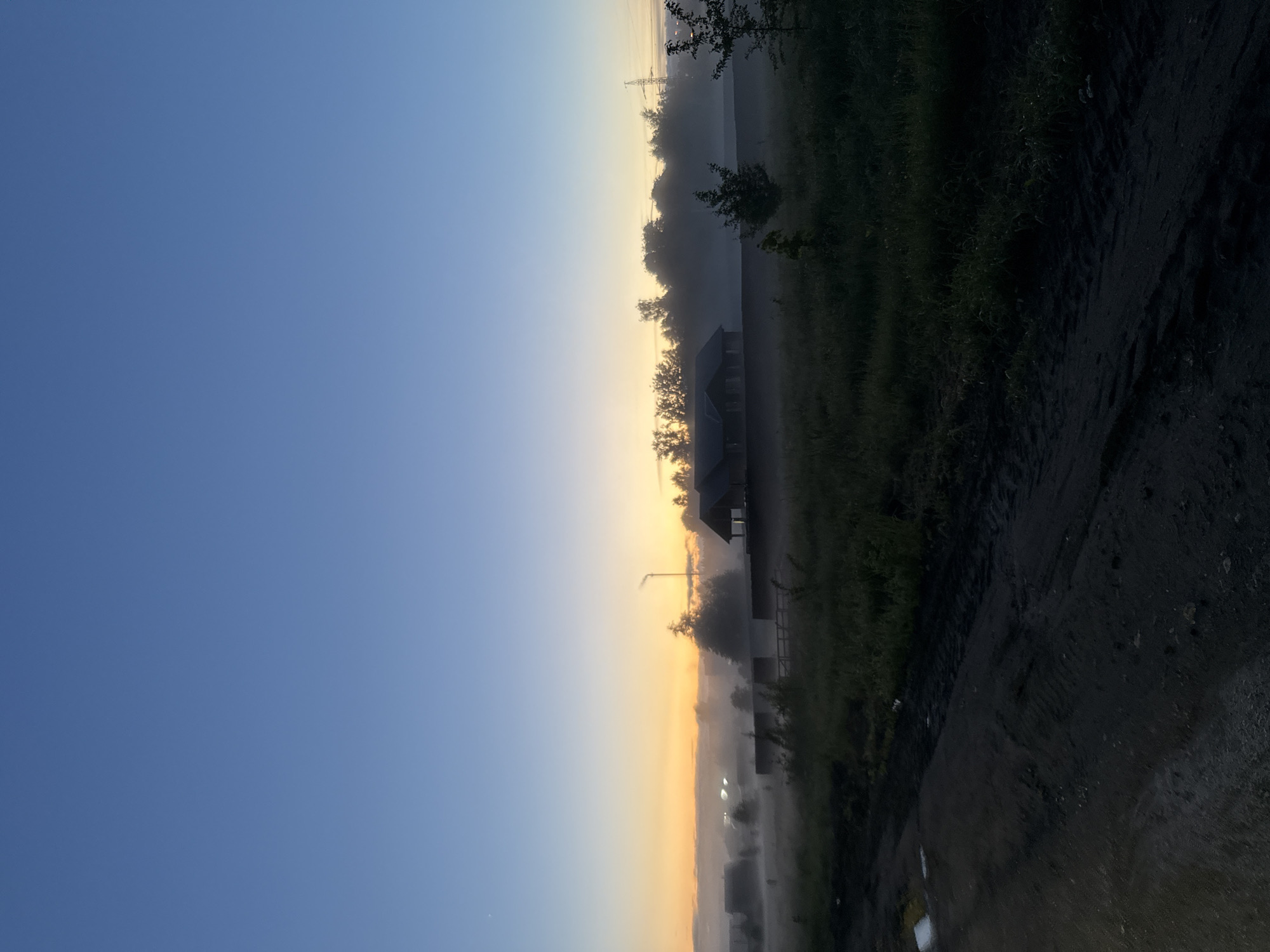 The image shows an outdoor scene in Buzhaninovo, Russia, at dawn or dusk. The sky transitions from dark blue to light yellow-orange near the horizon, indicating sunrise or sunset. Fog hangs in the air, obscuring details of the landscape. A house or building is visible in silhouette against the horizon. Trees and vegetation line the right side of the frame. A dirt road or path occupies the bottom right corner, with visible tire tracks and puddles. A street light is visible in the fog. Power lines are also visible in the distance. The lighting is dim and soft, with shadows cast by the low angle of the sun.
