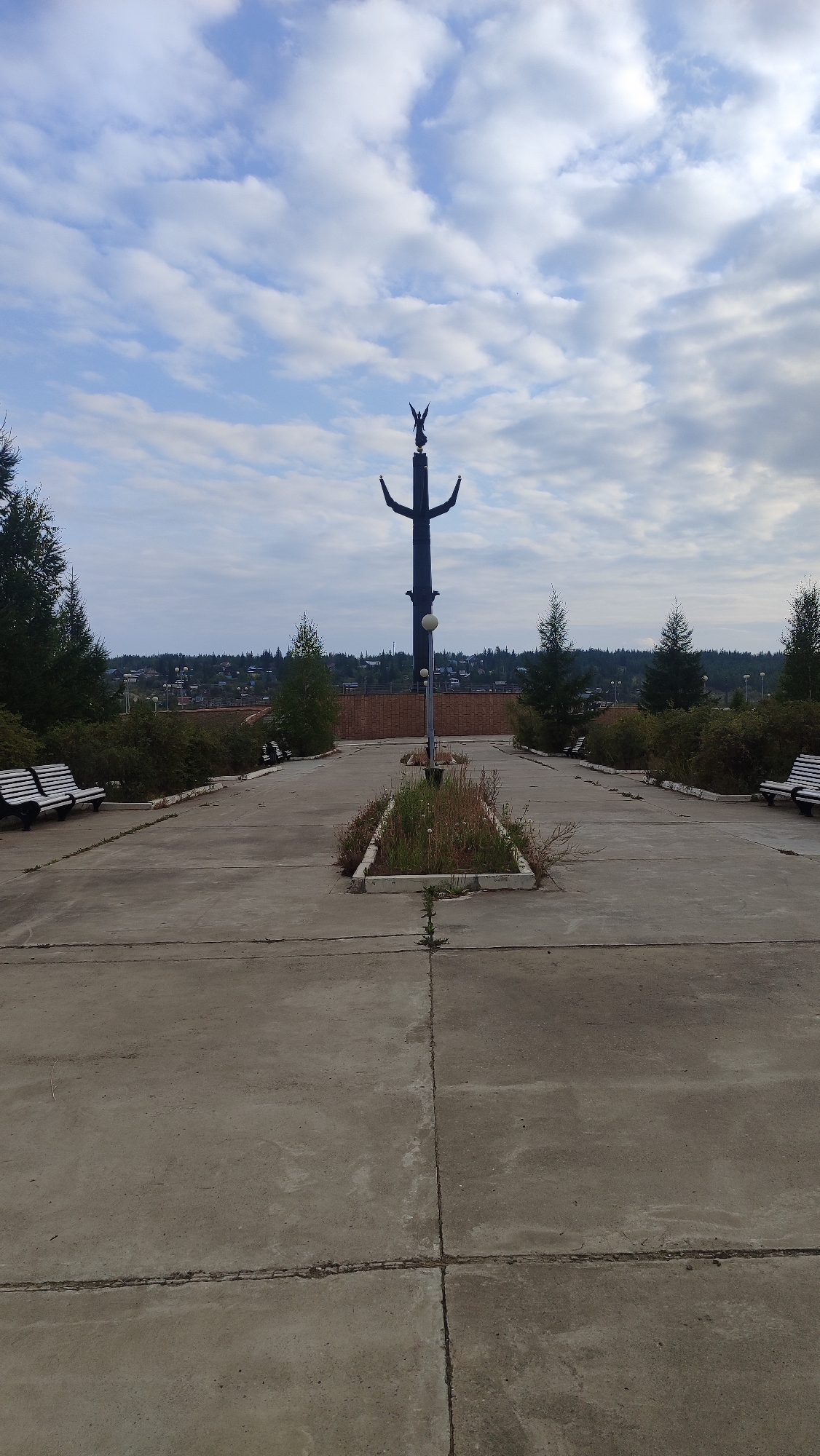 The image depicts a wide, concrete plaza with a central monument. The monument is a dark, slender structure with two curving arms near the top, and a small figure, possibly a bird or angel, perched atop it. Two white benches with dark slats are visible on either side of the plaza, near the foreground. Shrubs and small trees line the edges of the plaza, and beyond them, a line of coniferous trees separates the plaza from a town with buildings. The sky is filled with a mix of white and gray clouds. The location appears to be a public outdoor space, likely in a town or city, given the presence of the town in the background. It is likely an area designed for recreation or commemoration, with the monument and benches suggesting a place of significance. The overall atmosphere is calm and overcast. The image is taken from a slightly elevated position, looking towards the monument and the town beyond. This scene could be located in Mirny, Russia, a city known for its diamond mining history and located in the Sakha Republic.
