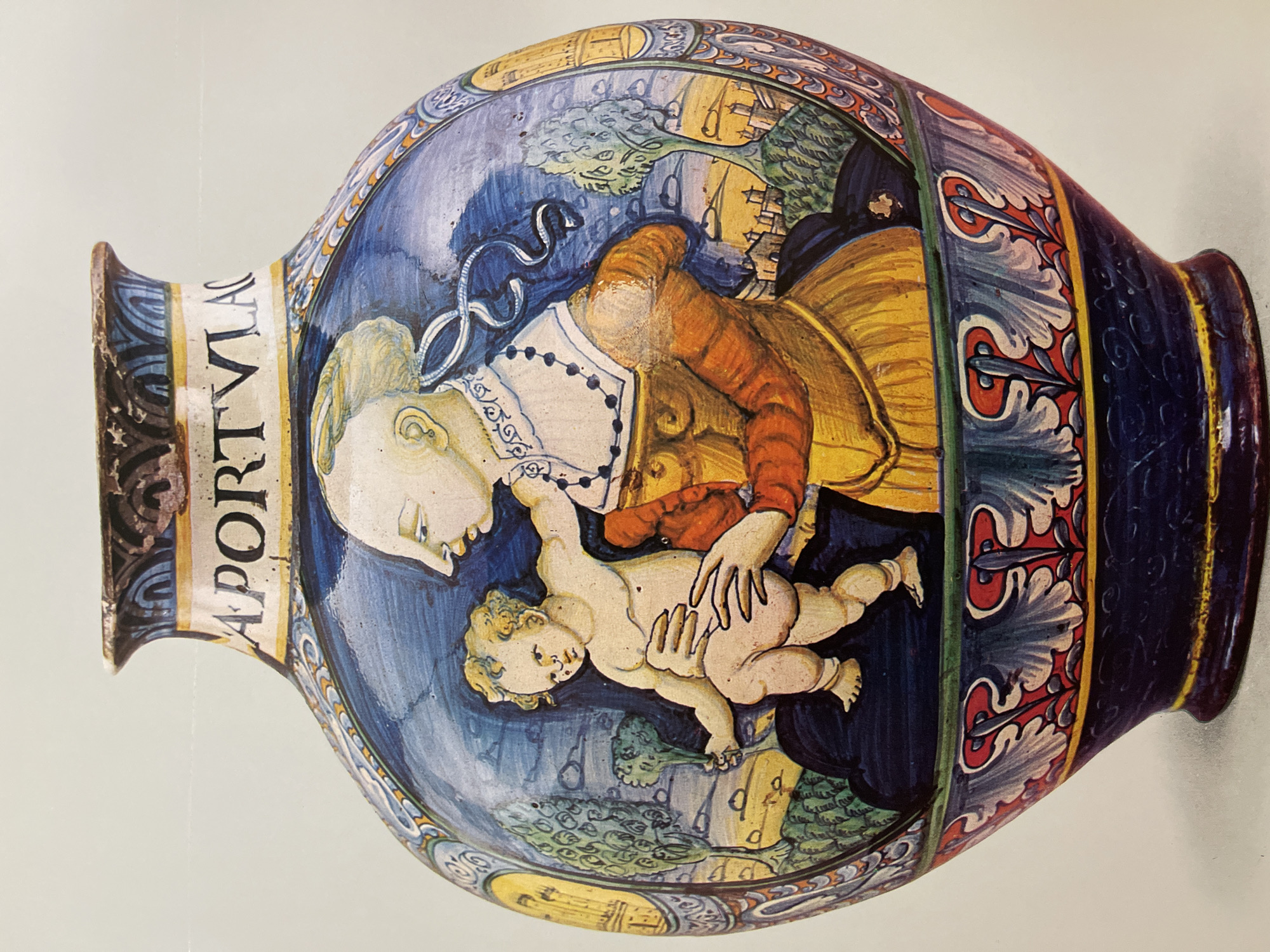 The image shows a large, decorative ceramic vase or urn. The main scene depicted on the body of the vase features a woman holding a baby. The woman has fair skin, blonde hair styled with ribbons, and is wearing a white-collared garment with a strand of beads and an orange and yellow gown. The baby is also fair-skinned. The background includes landscape elements such as trees and what appears to be a building in the distance. The vase is painted with blue and yellow colors, with ornate decorations at the top and bottom. There is text on the side of the vase near the top, running vertically. The photo does not appear to have been taken in Almaty, Kazakhstan.