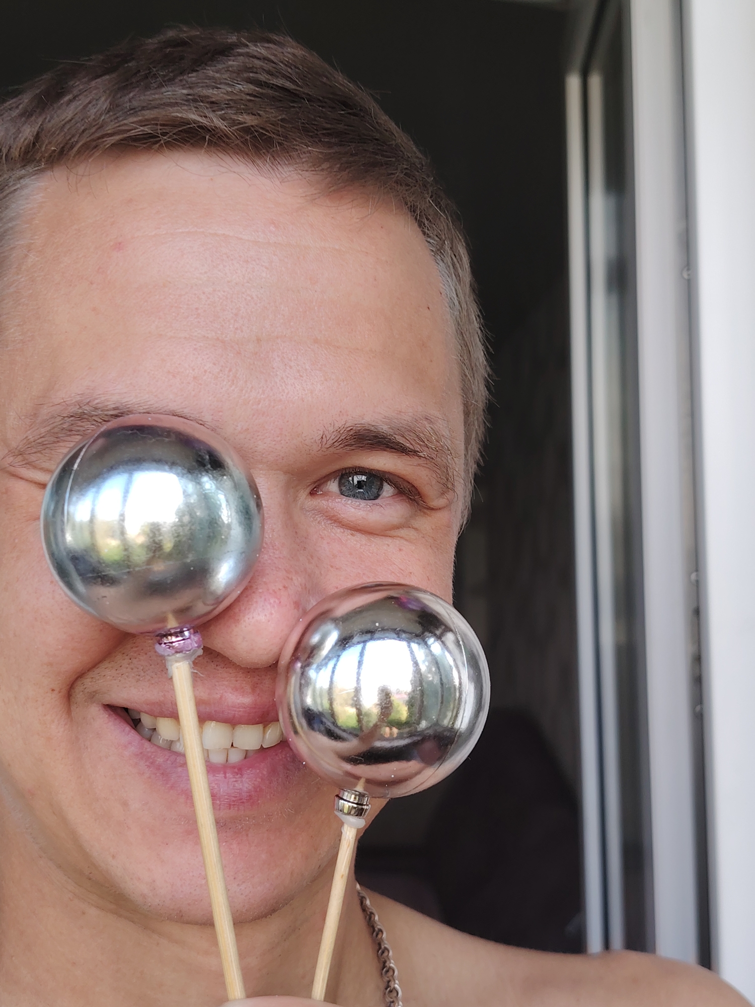 A close-up, low-angle, outdoor, medium shot shows the upper torso and face of a person, possibly male, who is smiling with their teeth showing. The person is holding two metallic, spherical ornaments on wooden sticks in front of their face, obscuring part of their left eye and nose. The left eye is visible and appears to be blue. The person has short, light brown hair with some graying at the temples. The background is out of focus and consists of a dark area to the left and a bright, vertical element to the right, possibly a window frame or doorway. The person is wearing a silver chain necklace. The scene suggests a casual, possibly playful or festive moment. The location is not identifiable from the visual information.