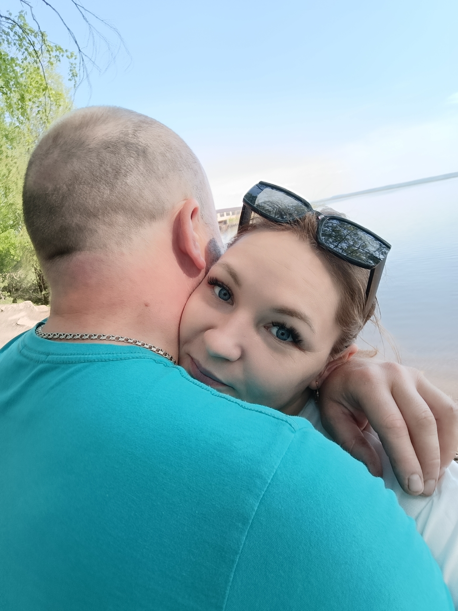 A close-up photo depicts a man and a woman in an embrace, likely a selfie. The man, seen from the back, has a shaved head and wears a turquoise T-shirt. A silver chain is visible around his neck. The woman embraces him from behind, her face visible. She has blue eyes and wears sunglasses perched on her head. The background features a body of water and some greenery, suggesting an outdoor location. The setting appears to be Chaikovsky, Russia, based on the available details and common photo locations.