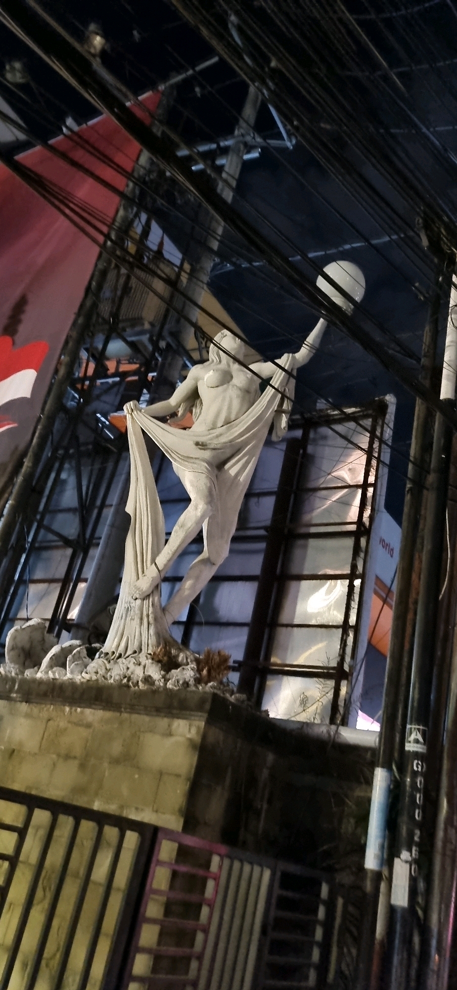 The image captures a nighttime view of a statue in Denpasar, Indonesia, likely a public artwork or monument. The statue depicts a female figure draped in flowing fabric, with her arms outstretched. Behind the statue, a large red and white banner, possibly the Indonesian flag, is visible, as are numerous overhead electrical wires and cables, suggesting an urban environment. A spherical light fixture can be seen in the upper right portion of the frame, illuminating the scene. The base of the statue rests on a stone platform, with a metal railing in the foreground. The background shows a building structure with some reflective surfaces and a partial view of what appears to be another banner with the word "world" visible. The overall impression is that of a city street or plaza at night, with the statue as the central focus, set against a backdrop of urban infrastructure and signage.