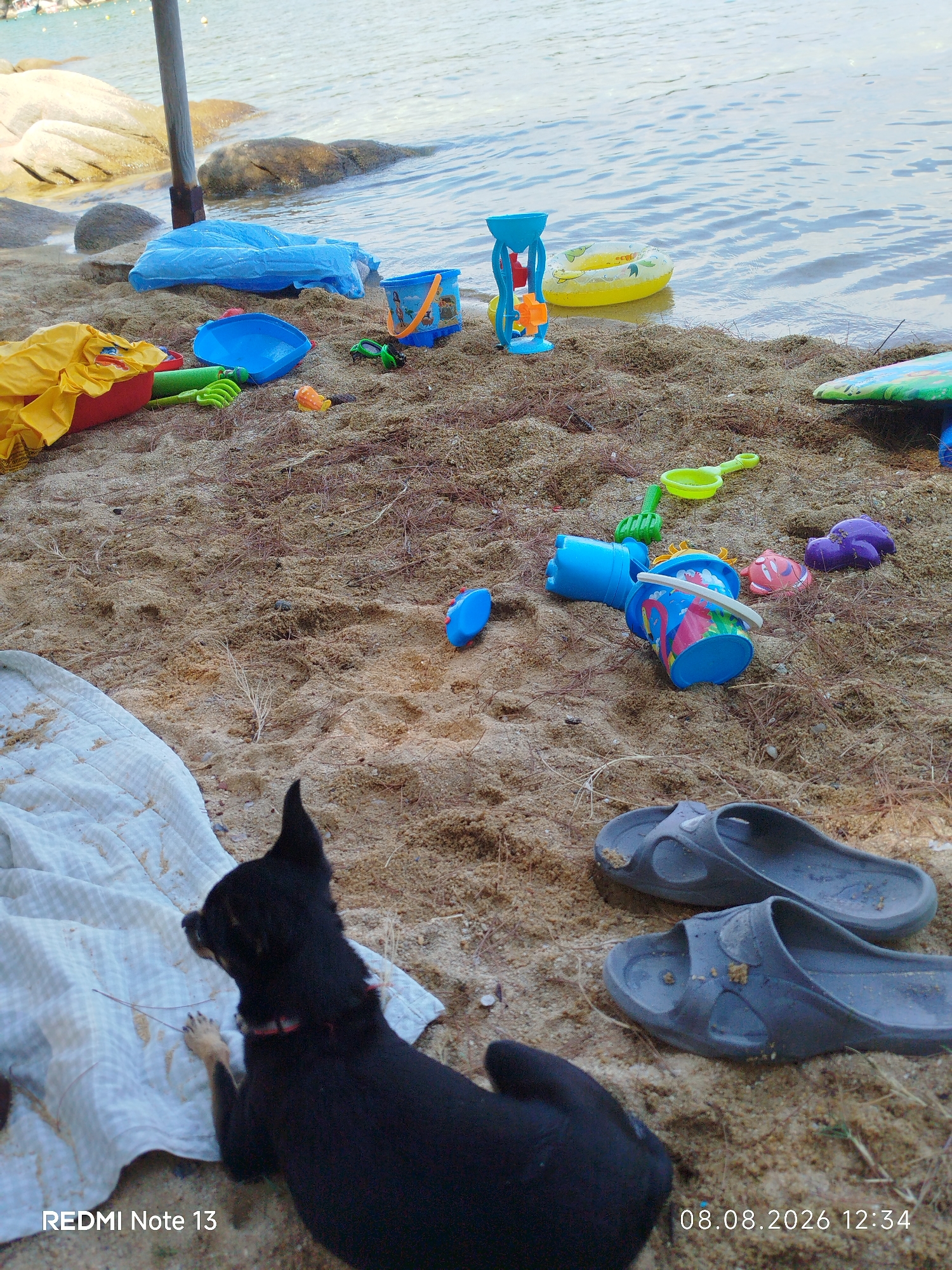 A black dog lies on a white blanket on a sandy beach in Sithonia Municipal Unit, Greece, with several children's beach toys scattered on the sand. The water of the sea is visible in the background, with large rocks on the left. The toys include buckets, shovels, a sand mold in the shape of a crab, and a small inflatable ring. A pair of gray flip-flops rests on the sand in the foreground. The timestamp on the image indicates it was taken on August 8, 2026, at 12:34.