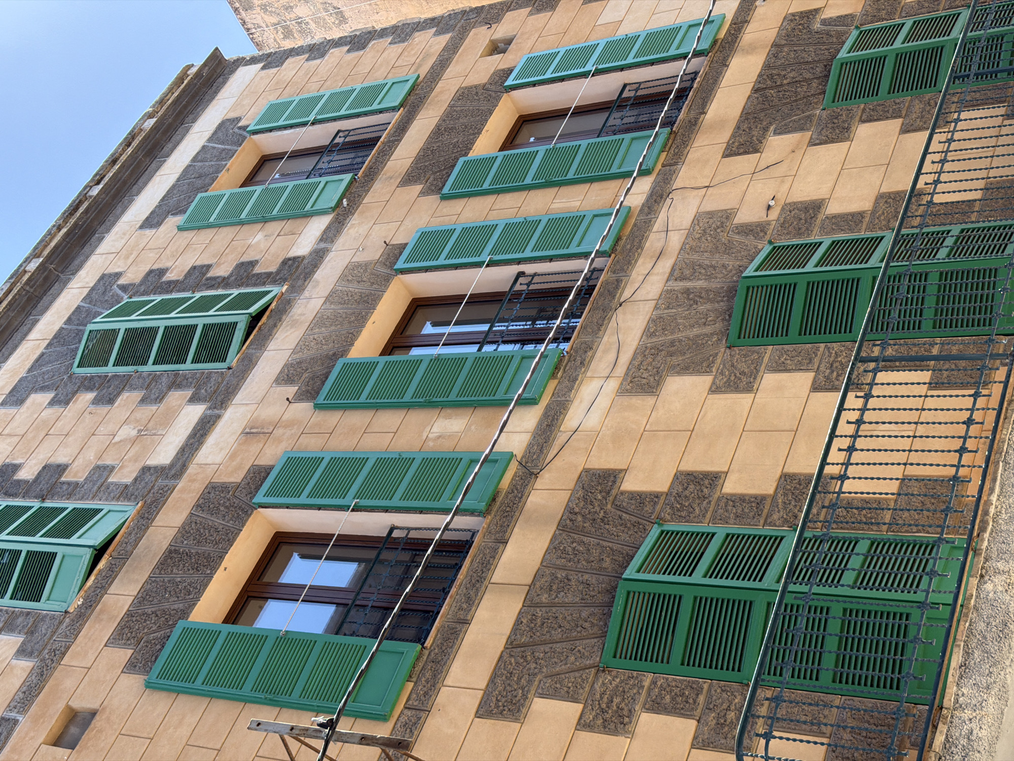The image shows the facade of a building in Manacor, Spain, featuring a repeating pattern of light brown and dark grey stone blocks. The building has multiple stories with numerous windows, many of which are adorned with vibrant green louvered shutters. Some shutters are open, revealing glimpses of windows and dark railings on small balconies. Wires run horizontally across the facade. The sky above is a clear blue. The architecture suggests a traditional style common in Mediterranean regions.