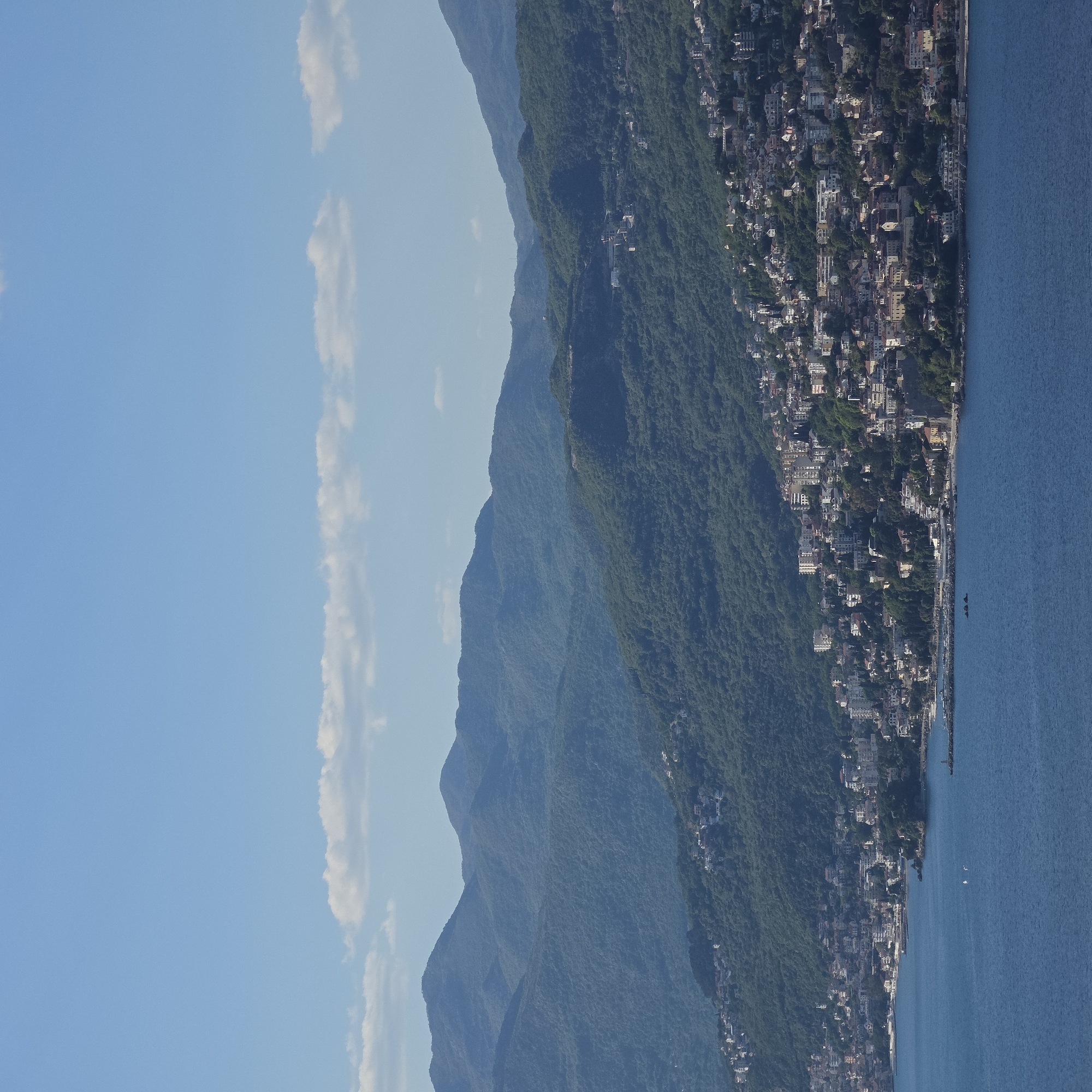 The image presents a panoramic view of a coastal landscape. In the foreground, a calm, deep blue sea stretches across the bottom portion of the frame (when oriented correctly). Along the shoreline, a town or settlement is densely packed, featuring numerous buildings of various sizes and colors, nestled against the base of the mountains. Some structures appear to extend into the water, possibly as small piers or jetties.
The mid-ground and background are dominated by a series of majestic mountains, heavily covered in lush green forest. These mountains recede into the distance, with their contours becoming softer and hazier, illustrating atmospheric perspective. The sky, occupying the top portion of the image, is a clear, bright blue, accented by a few scattered, wispy white clouds.
No specific human activity is clearly discernible due to the wide perspective and distance; the scene appears peaceful and static. A few tiny, indistinct shapes on the water might be boats, but this is not certain.
The notable details include the bright daylight, indicated by the clear blue sky and strong illumination, suggesting either morning or afternoon. The weather is fair and clear. The landscape, with its combination of steep, forested mountains, a coastal town, and the serene sea, strongly aligns with the geographical characteristics of Luštica, Montenegro, a region known for its rugged coastlines and picturesque settlements. The overall mood is one of tranquility and natural beauty.
There is no visible text in the image.