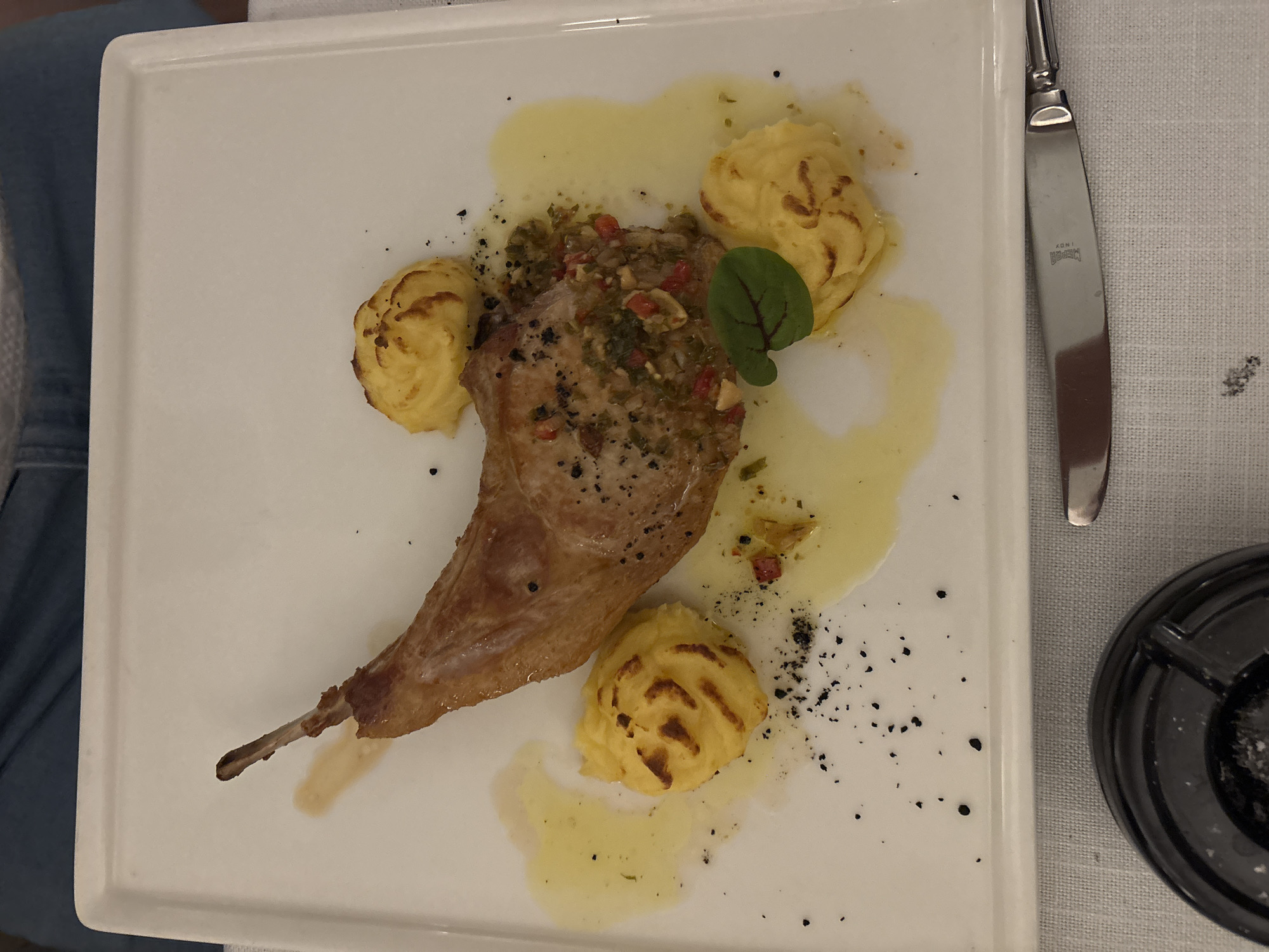 A white plate of food is presented on a white tablecloth, with a person's legs in blue jeans visible in the upper background. The plate contains a portion of roasted meat with a bone, served with three dollops of mashed potatoes piped in a spiral pattern. The meat is garnished with a chopped relish containing herbs and red pepper flakes, and drizzled with a light yellow sauce, possibly butter or oil. Black specks, likely pepper or ground spices, are scattered across the plate and tablecloth. A small green leaf garnishes the dish. A knife with a polished metal handle and blade is positioned on the right side of the plate. An ashtray is visible in the lower left corner of the image. The scene is indoors, likely in a restaurant in Varna, Bulgaria.