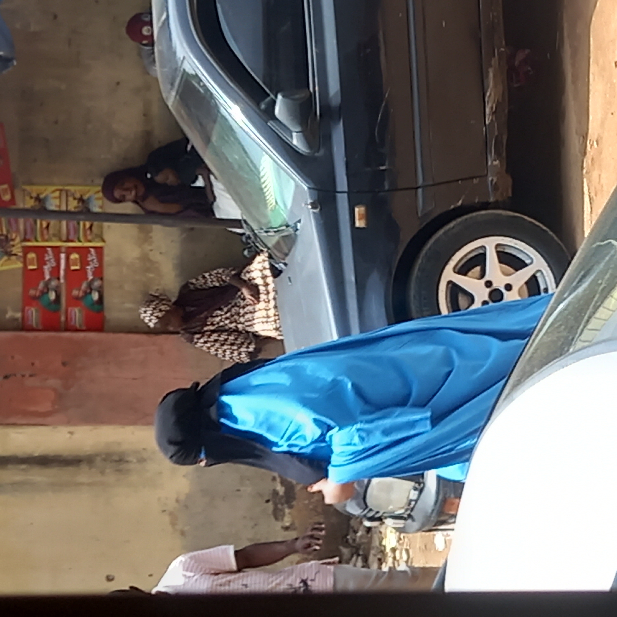 The image, rotated clockwise, depicts a scene in Dadi Riba, Nigeria. A dark-colored car is parked on a dirt surface near a wall. Adjacent to the car, individuals are present. One person wears a long blue garment with a black niqab covering their face. Beside them, an individual wearing a light pink, striped shirt is visible. Behind the car, there are two women seated. One of the women is wearing a patterned brown and white outfit, and the other one is wearing a dark colored outfit. Further behind them is what appears to be advertisement posters on a wall.