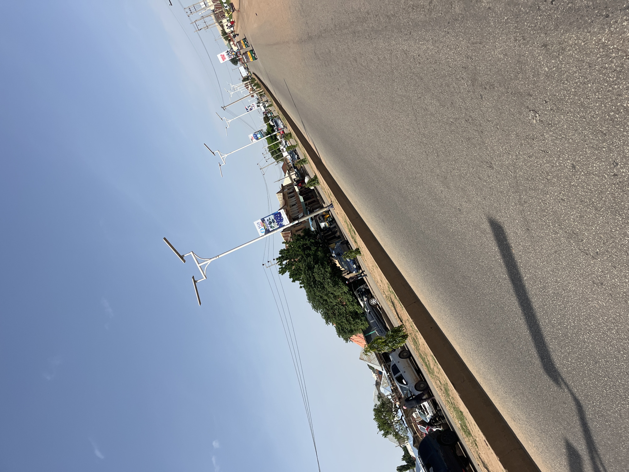 The image shows a street scene in Bukuru, Nigeria. A paved road runs diagonally from the bottom right to the top left of the frame. On the left side of the road, there is a sidewalk with a low brown curb. Behind the sidewalk, there are several buildings with corrugated metal roofs, some of which appear to be shops or residences. Trees and other vegetation are visible between the buildings and along the sidewalk. A line of streetlights with solar panels is positioned along the sidewalk.
Several vehicles are parked along the side of the road or on the sidewalk, including a black SUV on the far left, a silver sedan, and several other cars. A few people are visible walking on the sidewalk or near the vehicles. In the background, more buildings and trees can be seen, along with power lines and utility poles.
Further down the road, traffic is visible, including a yellow and green motorized tricycle. Several billboards or signs are also present, some with text and graphics. The sky is clear and blue with a few wispy clouds. The overall impression is of a busy street in a Nigerian city.
