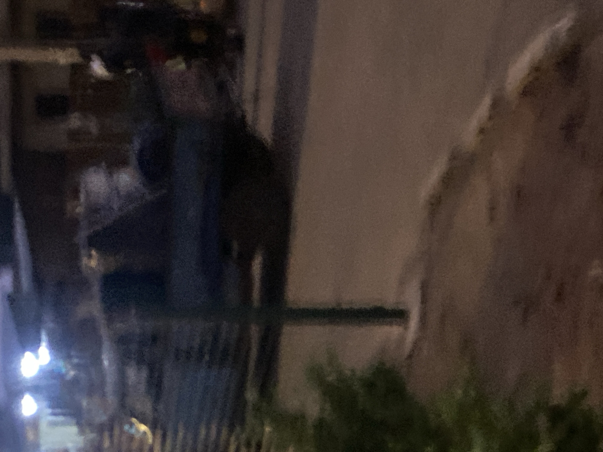 The provided image captures an outdoor street scene at night or late dusk, identified as being in the city of Bukuru, Nigeria. The photograph exhibits significant motion blur and high digital noise, resulting in poor clarity and indistinct details.
In the foreground, a vibrant green bush occupies the lower-left corner. A dark green pole, possibly a utility or signpost, stands vertically from the lower-center extending upwards, positioned near the edge of a paved surface.
The central part of the image depicts a dark-colored road surface, likely asphalt or concrete, extending from the lower-center towards the background. On the left side of the road, a dark, indistinct vehicle, possibly a pickup truck or utility vehicle, is present, facing generally towards the right. Its outline is blurry, but a cargo bed or rear section is discernible, possibly containing dark, bulky items. Several bright, white-blue artificial light sources are visible in the upper-left quadrant, consistent with streetlights or distant vehicle lights.
On the right side of the road, a concrete curb line separates the roadway from an elevated, textured area, possibly a sidewalk or verge. Adjacent to this curb, on the right side of the frame, a person is visible next to what appears to be a motorcycle or scooter. The individual is wearing dark clothing and a light-colored helmet. Hints of red and yellow are present on or near the motorcycle/person, suggesting vehicle coloration or accessories. The person's posture is unclear, but they appear to be stationary or moving slowly.
The background consists of dark, indistinct structural outlines, likely buildings, under dim ambient lighting. No specific identifying features of these structures are discernable due to the image's blurriness and low light conditions. The overall color palette is dominated by dark blues, greens, and browns, with bright white highlights from artificial light sources.
