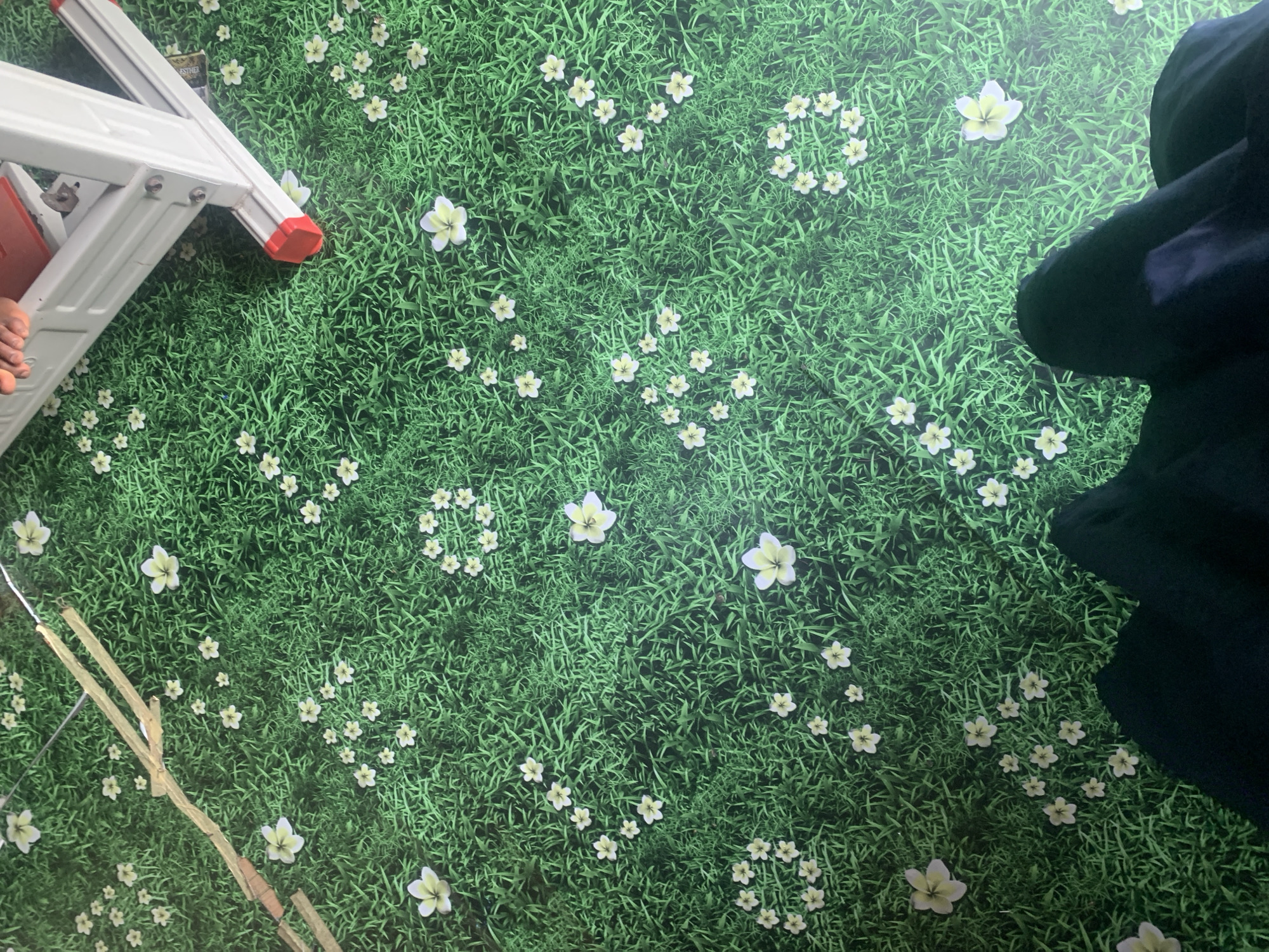 An overhead shot captures a patch of green grass with scattered white flowers, resembling a stylized lawn or carpet. Two wooden planks lie diagonally across the upper left portion of the frame, appearing to be part of a construction or decorative element. In the upper right, the white and red legs of a ladder are visible, with a person's feet peeking over the top. The bottom of the image shows dark fabric, likely clothing. The scene is devoid of any identifiable urban or rural features of Jauro Yinu, Nigeria.