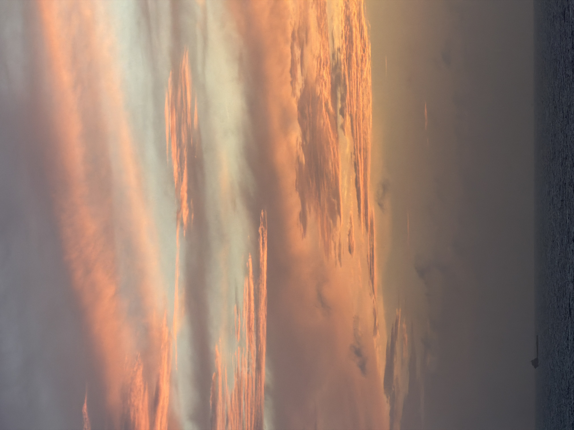 The image shows a view of the sky at sunset, with elongated, vertically-oriented clouds tinged with orange and pink hues. The left side of the image features lighter, diffused areas while the right side transitions to darker, more concentrated cloud formations. A sliver of what appears to be the ocean or a dark body of water is visible on the far right edge. The location in the city is unknown, Unknown.