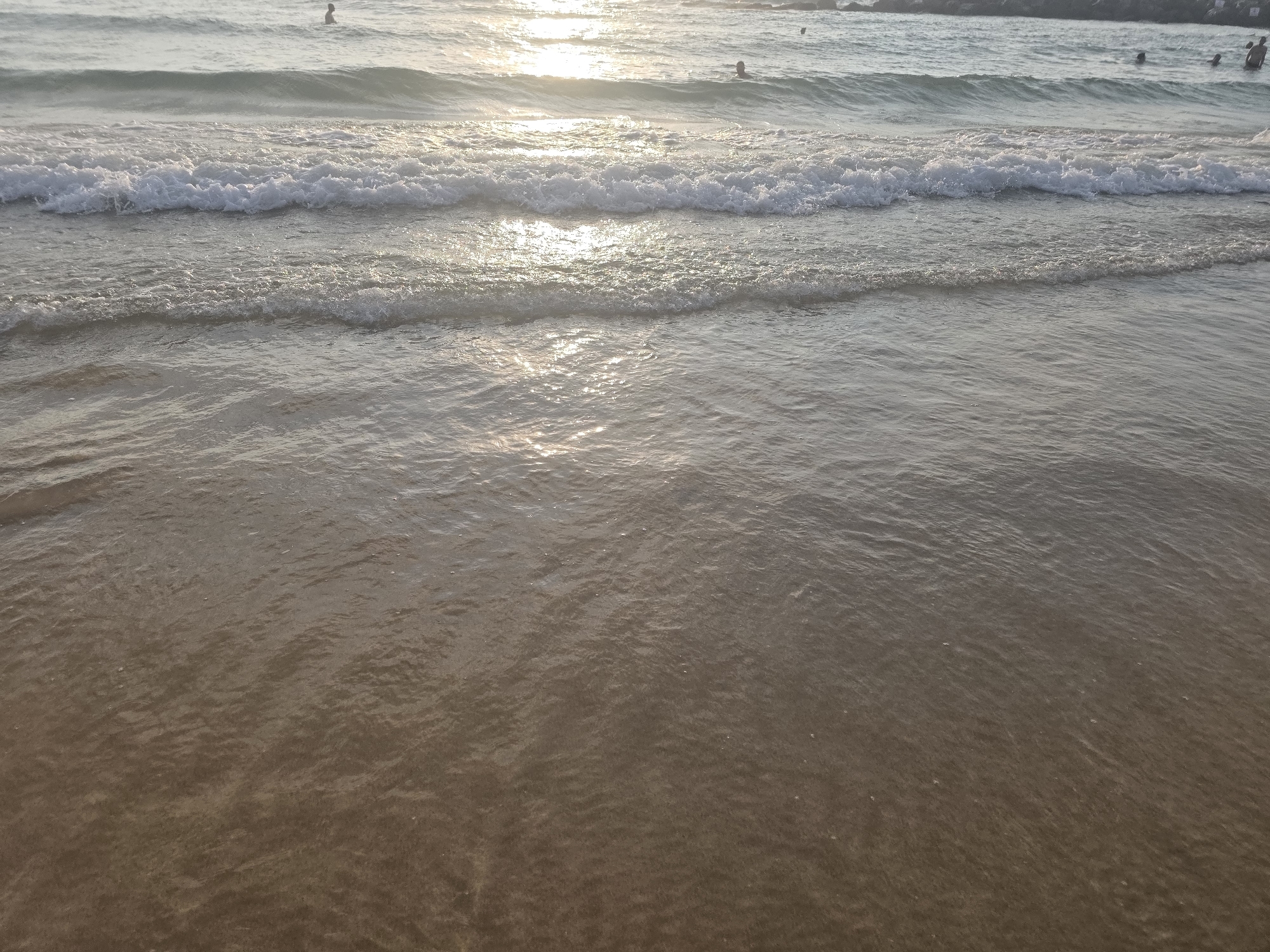The image depicts a beach scene at dusk in Ashkelon, Israel. The foreground is dominated by shallow, clear water with a sandy bottom, reflecting the soft, golden light of the setting sun. Small waves gently lap at the shore, creating ripples and subtle patterns on the water's surface. In the middle ground, the ocean stretches out, with several lines of breaking waves in varying sizes, their white foam contrasting with the darker water. The setting sun casts a bright, shimmering reflection across the water's surface. In the distance, beyond the breaking waves, a few individuals are visible in the water, their figures silhouetted against the bright sky. The shoreline in the far background is indistinct, appearing as a darker line against the horizon, possibly with some rocky outcroppings. The overall atmosphere is peaceful and serene, characteristic of a beach at the end of the day.