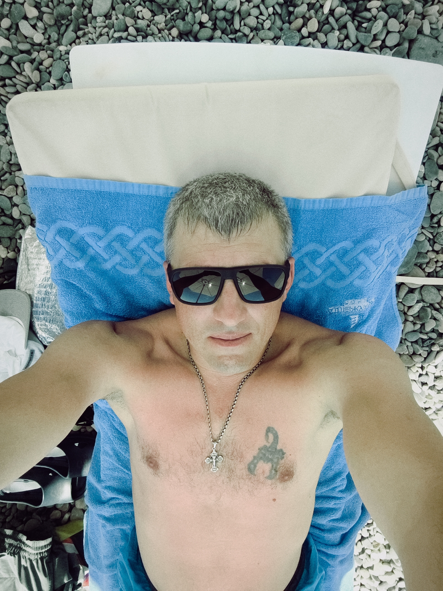 The media file shows a man lying on a blue towel and a white pillow on a pebble beach. He is wearing sunglasses and a silver chain with a cross pendant. He has a tattoo of a scorpion on his chest. The background is filled with gray and white pebbles. The scene appears to be set outdoors during the daytime, with the sun casting a warm glow on the man's skin, suggesting good weather. The man's expression is relaxed, and he is looking directly at the camera, possibly taking a selfie. The location context is Sochi, Russia. There is visible text on the blue towel, but it is not clear enough to be read.