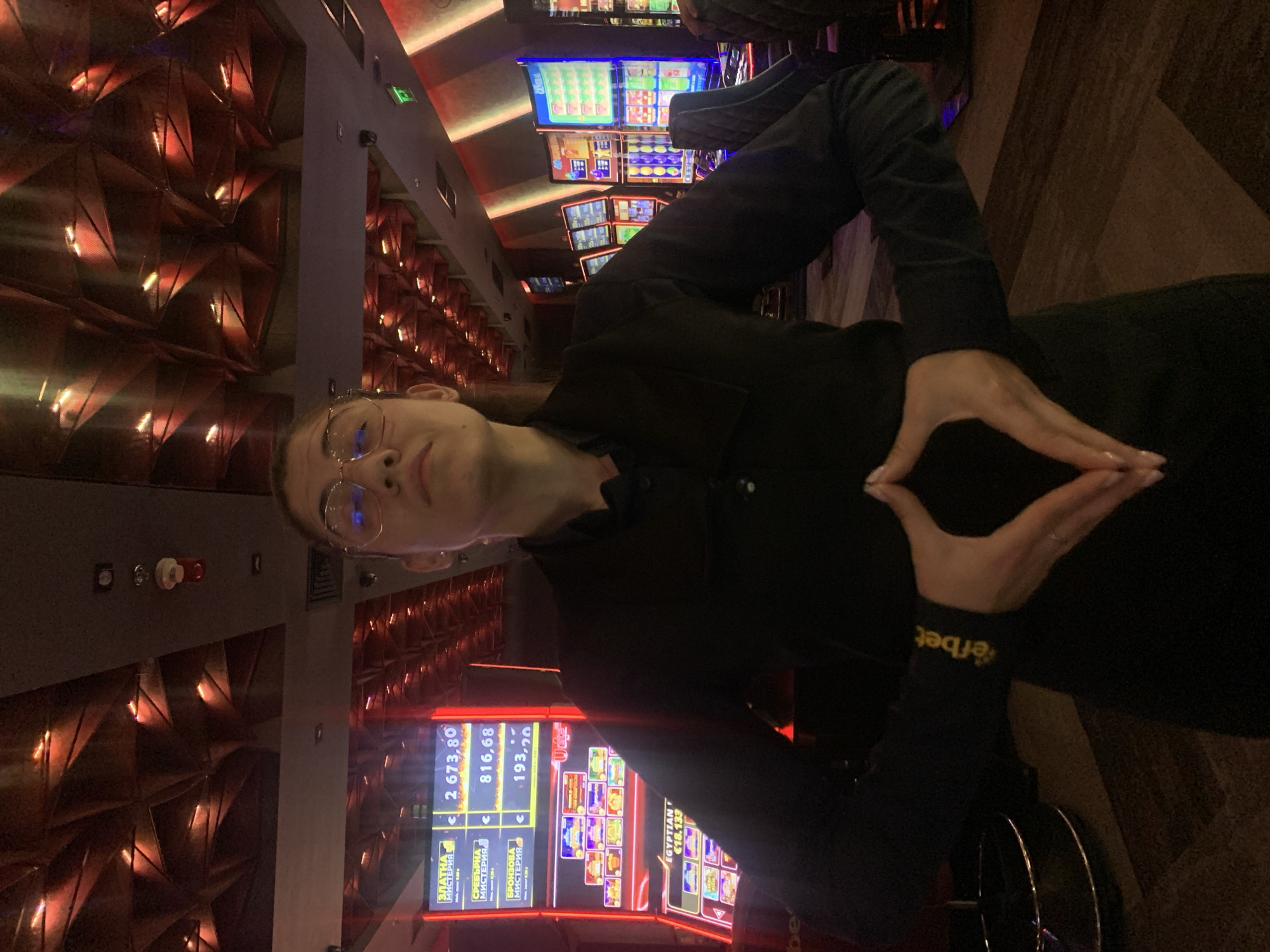 The image shows a woman in a black shirt and glasses, standing in what appears to be a casino. Her hands are positioned in front of her chest, forming a diamond shape. Behind her, there are rows of slot machines, some of which are lit up. The ceiling has an intricate design with red lighting. The woman's shirt has a yellow logo on the left sleeve that reads "efbet". This logo is associated with Efbet, a Bulgarian betting and casino company, which has multiple locations in Burgas, Bulgaria.