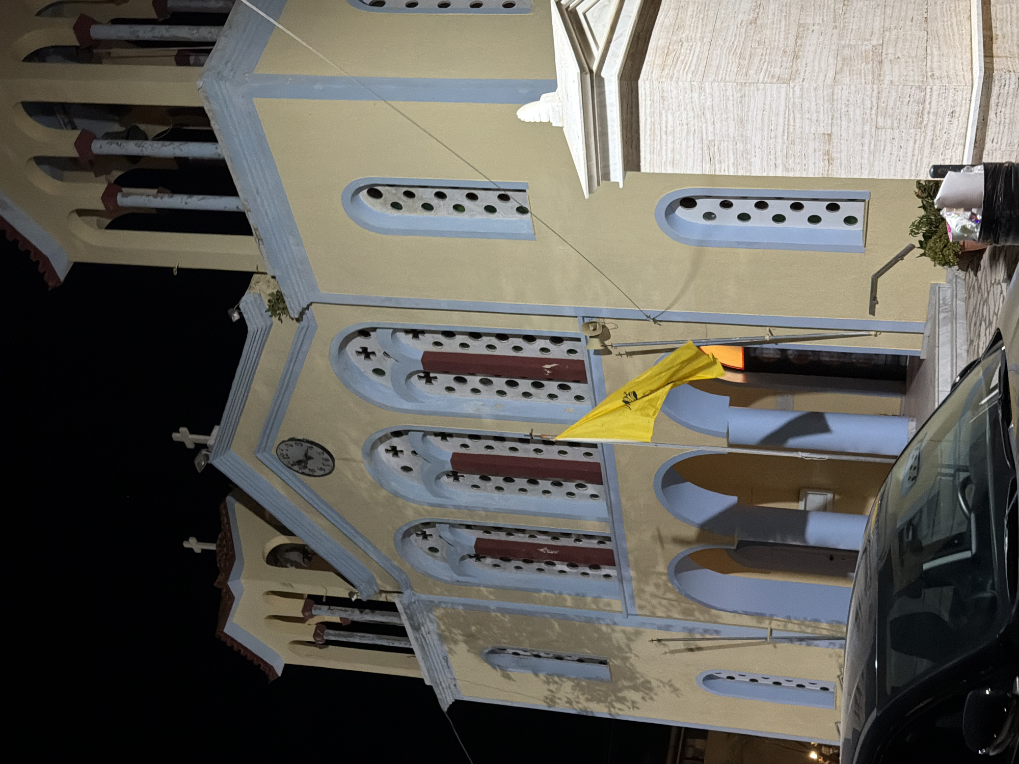 The image captures a nighttime view of a church facade in the Municipal Unit of Alyzia, Greece. The church is painted in a pale yellow with blue trim around the arched windows and doorways. Several tall, arched windows with intricate latticework and red interior shutters are visible on the main facade. A clock face is positioned above the central arched entrance. A yellow flag, possibly religious in nature, hangs from a pole to the right of the entrance. The church features bell towers with arched openings. In the foreground, the roof and windshield of a black car are visible. The environment is dimly lit by artificial light, casting shadows. The ground is paved with cobblestones. A modern marble-clad structure with a decorative cap stands to the right of the church.