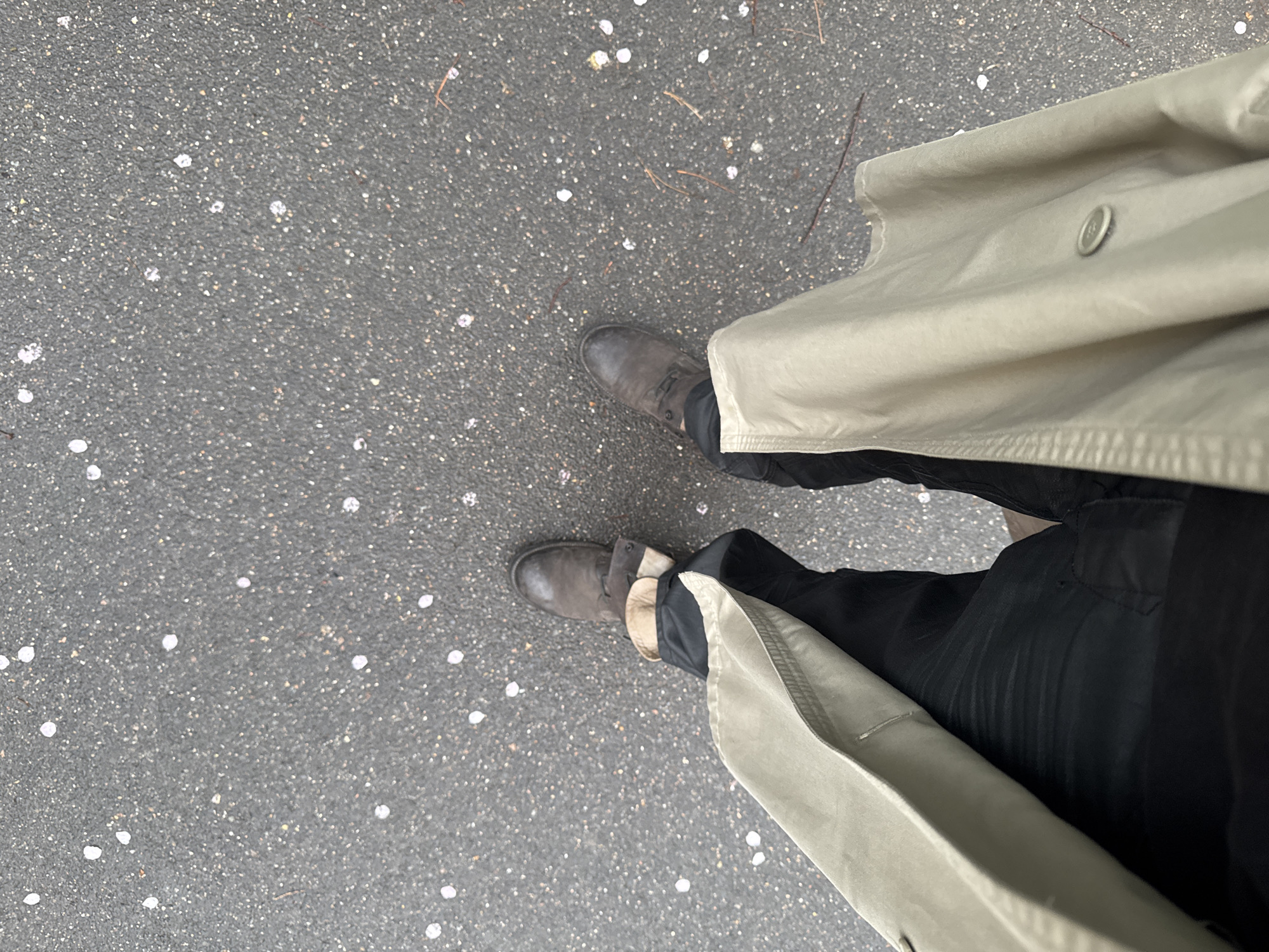 A person stands on an asphalt surface scattered with small white spots, possibly flower petals, indicative of springtime. The individual wears dark trousers and brown leather boots. A light-colored trench coat, buttoned at the top, is draped around them. The location is likely an outdoor area in Les Ulis, France, based on image context.