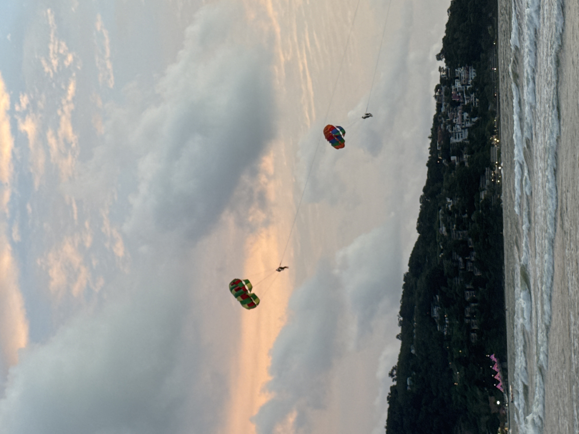 In Patong, Thailand, during sunset, two colorful parasails are visible against a dusky sky filled with soft pink and grey clouds. One parasail, colored green and red, is higher and further to the left, with a single person suspended beneath it. To the right and lower, a second parasail, with red, blue, and green sections, has two individuals attached. Both parasails are tethered to an unseen tow boat, their lines stretching taut across the frame. Below the sky, a densely populated hillside is illuminated with scattered lights, suggesting residential buildings nestled amongst dark green foliage. The foreground features a choppy ocean with white-capped waves rolling towards the shore.
