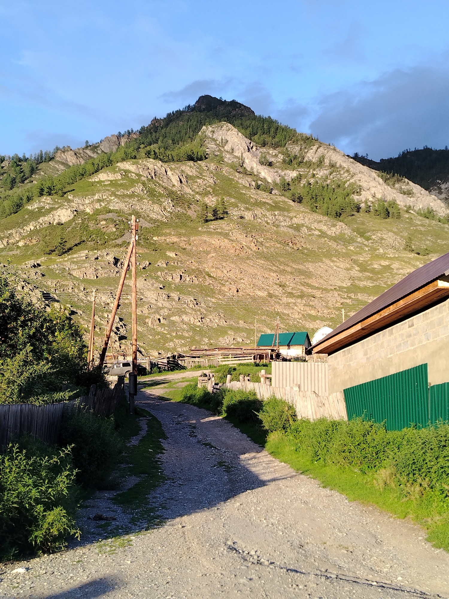 The image shows a dirt road leading towards a mountain in Iodro, Russia. The road is lined with fences and lush green vegetation. On the right side of the road, a building with a metal roof and a concrete block wall is visible. Further down the road, there are smaller structures, including one with a green roof. A utility pole stands tall on the left side of the road. The mountain in the background is covered with trees and rocky outcrops. The sky above is blue with some scattered clouds. The overall scene depicts a rural setting with elements of both natural landscape and human habitation.