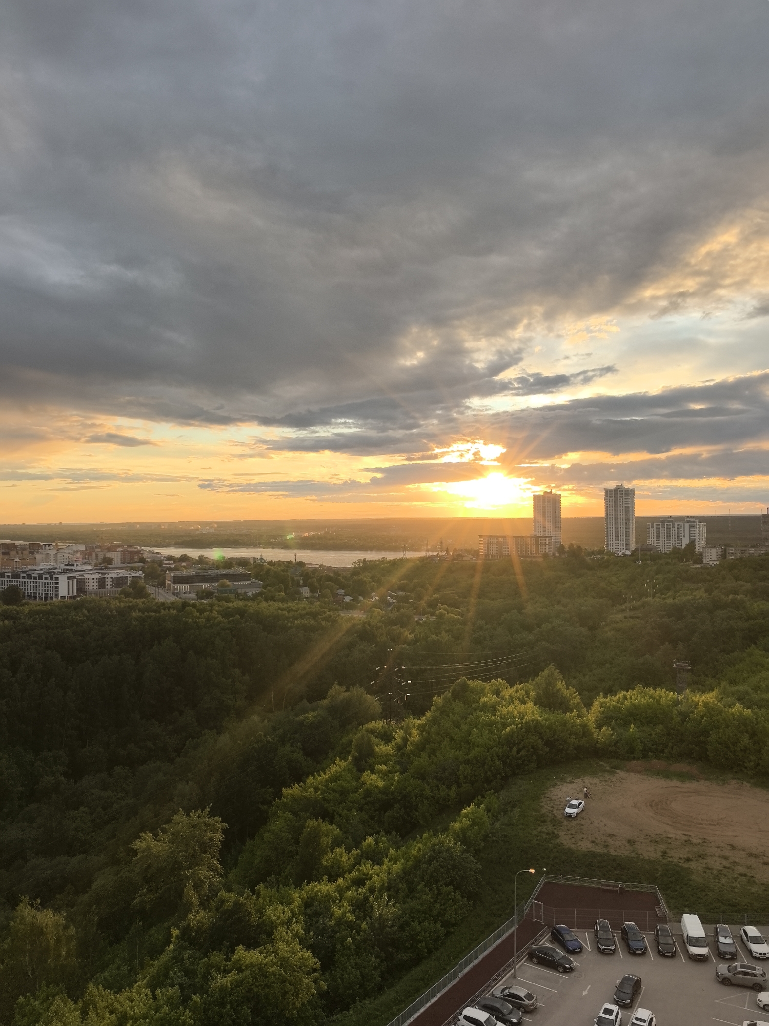 The image is a wide, elevated view of Perm, Russia at sunset. The sky is filled with layers of grey and orange clouds, with the sun breaking through the clouds on the horizon. The sun's rays are prominent, casting light on the landscape below. In the mid-ground, there are several tall buildings. In the foreground, dense green trees cover the landscape, partially obscuring a flat dirt surface, with a white car on it and two people near the car. At the very bottom of the frame, there is a parking lot with multiple parked vehicles. A lamppost is located near the parking lot.