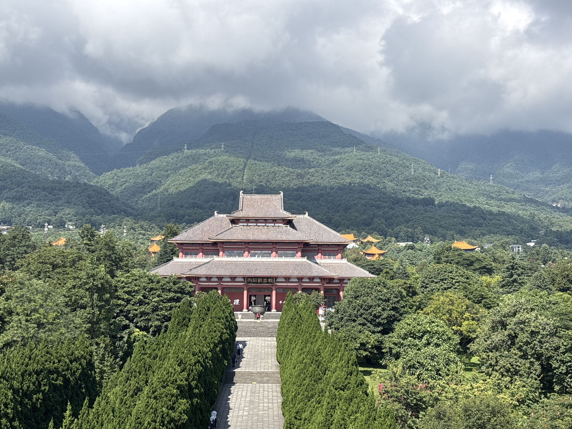 An aerial perspective captures the iconic Three Pagodas of Chongsheng Temple in Dali City, China. The central and largest pagoda, built during the Tang Dynasty, stands prominently, its intricate eaves and weathered facade a testament to centuries of history. Flanking it are two smaller pagodas, also of impressive age, completing the sacred ensemble. The pagodas are nestled amidst lush, verdant trees, their dense foliage creating a striking contrast with the ancient stone structures. In the background, the majestic Cangshan Mountains rise, their slopes shrouded in a mystical mist, adding to the serene and timeless atmosphere of the scene. A pathway, lined with manicured cypress trees, leads towards the main pagoda, inviting visitors to explore the spiritual grounds. The sky above is a canvas of dramatic clouds, some heavy with rain, others tinged with sunlight, creating a dynamic and atmospheric backdrop.