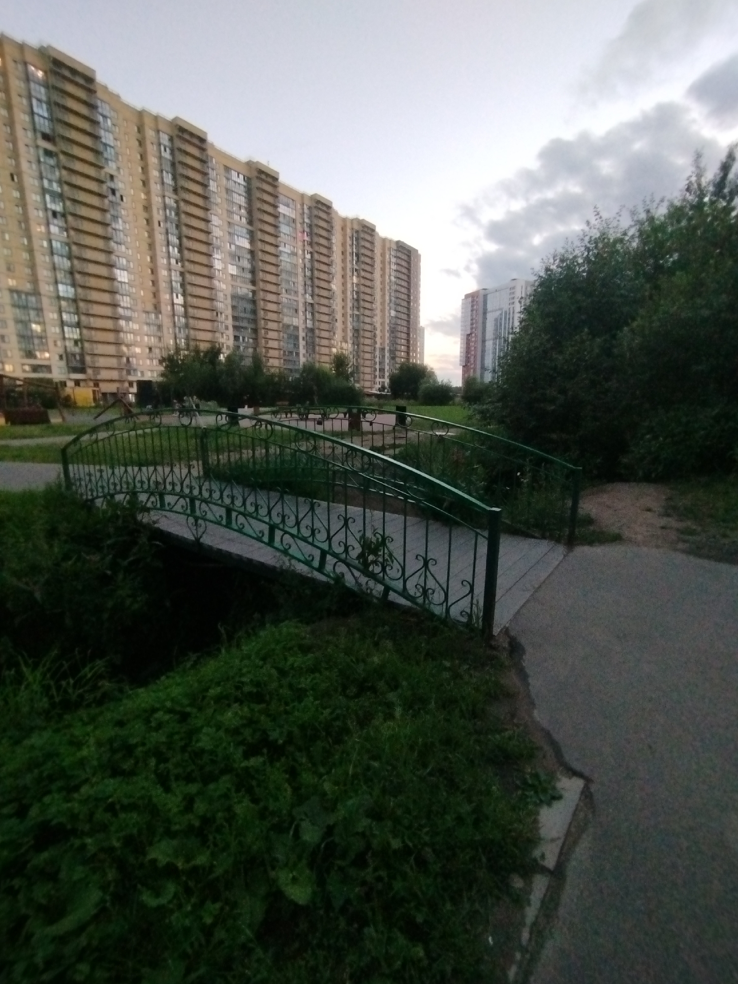 The image depicts a park-like area in the city of Pargolovo, Russia, with a focus on a decorative green metal bridge spanning a small water feature. Tall, modern residential buildings with numerous windows dominate the background, suggesting an urban or suburban setting. Lush green grass and trees surround the bridge and the pathway leading up to it. The sky is overcast with clouds, indicating it might be dusk or a cloudy day. No people or animals are visible in the scene.
