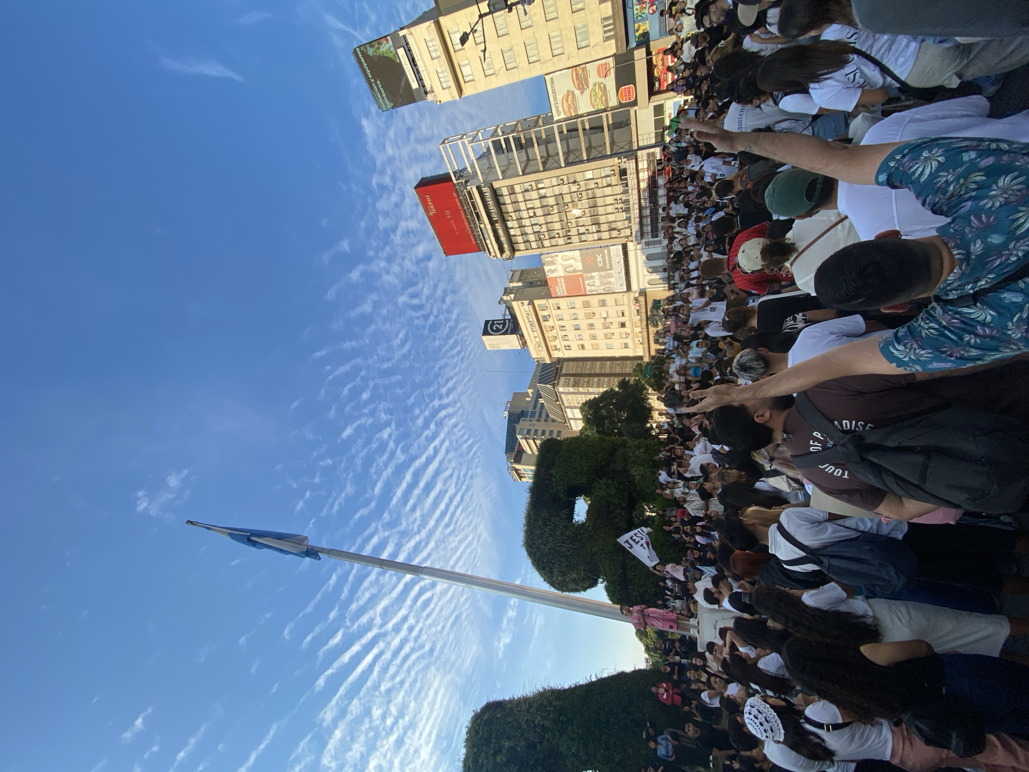The image captures a large gathering of people in Buenos Aires, Argentina. Buildings with advertisements, including those for Burger King, ICBC, and "formación y equipamie bomberos," line the street. A tall flagpole with an Argentinian flag stands amidst green bushes. The sky is blue with scattered clouds. The crowd appears to be facing the buildings, and a few hold signs, one of which reads "JESU T" and another reads "Tour OF P ADISE." People are wearing various clothing, and some have their arms raised.