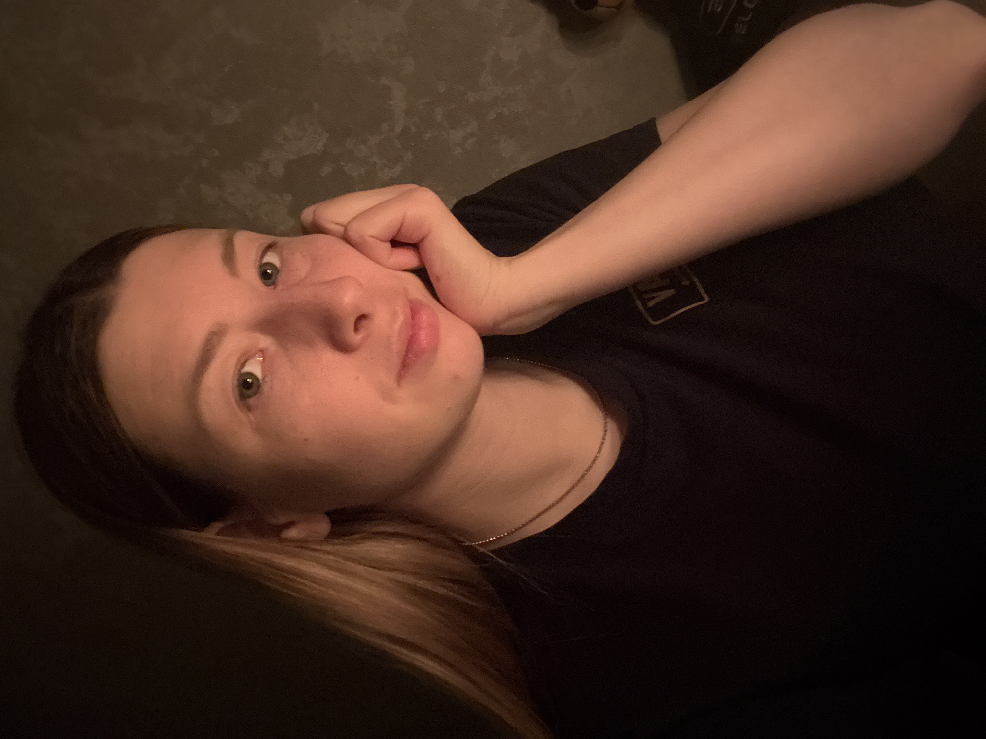 A close-up, dimly lit portrait features a young adult, possibly female, reclining indoors. Her head is tilted, and her left hand is casually positioned by her face, supporting her head. She looks towards the viewer with blue-green eyes and a neutral, relaxed expression, hinting at a soft, subtle smile. She has light skin and light brown hair with blonde streaks, which is styled down around her shoulders.
She is wearing a dark, short-sleeved t-shirt with a visible yellow logo that reads "VANS" on her left chest area. A delicate, thin metallic necklace is worn around her neck. The background is dark and features a textured surface, possibly a wall or furniture upholstery. In the upper right corner, an indistinct object with partial text, possibly "ELO" or "ELC," is barely visible. The lighting is low and warm, suggesting an evening or nighttime setting. The image conveys a casual and intimate mood. The location context for this image is Moscow, Russia.