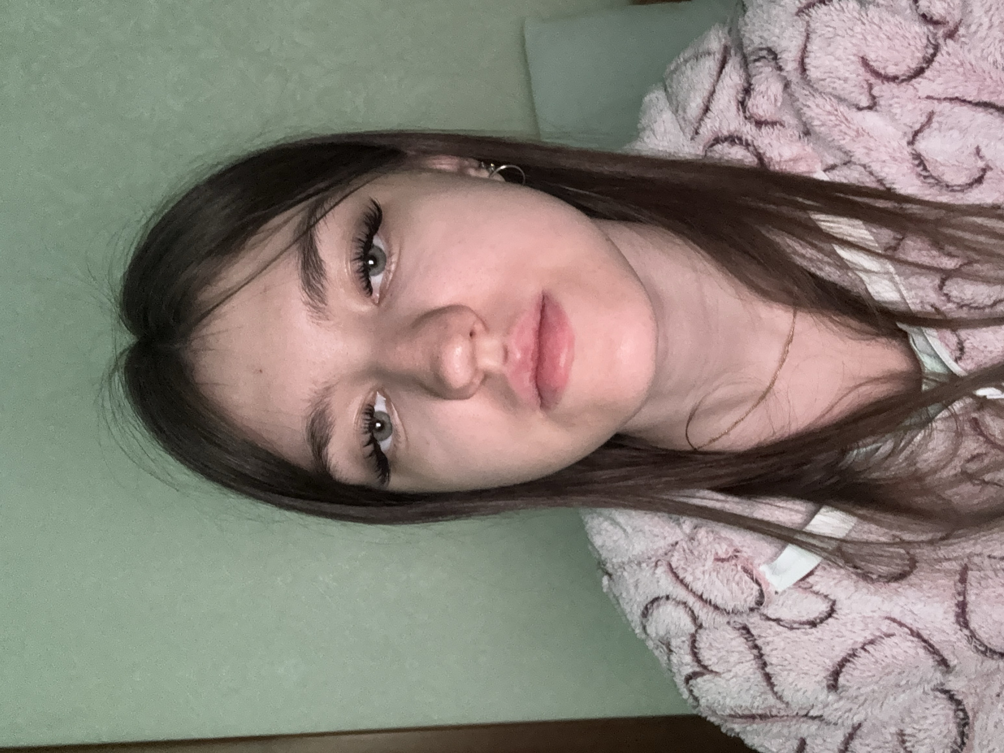 A young woman is captured in a close-up portrait, reclining indoors. She has long, dark brown hair, fair skin, and light-colored eyes, possibly grey-green, framed by long, dark eyelashes. Her lips are full and slightly parted, with a neutral to relaxed expression. She is wearing a small, silver-toned hoop earring in her visible ear and a thin, golden chain necklace is visible around her neck.
She appears to be wearing or covered by a fluffy, light pink robe or blanket adorned with a darker, swirled or abstract pattern. The background consists primarily of a light, muted green wall, with a strip of dark brown along the bottom edge, possibly a wooden trim or furniture. The lighting is soft and appears to be artificial or low light, suggesting an indoor setting during the evening or night. No discernible text is visible.