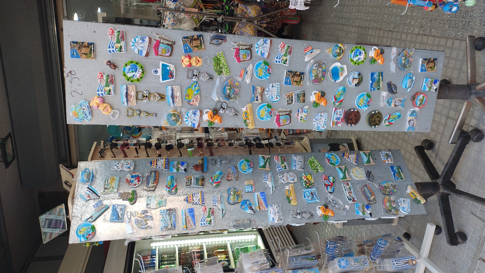 The image shows two tall, grey, rotating display stands filled with numerous colorful souvenir magnets. The stands are positioned outdoors, with a tiled ground surface visible and some shelving units with items on the left. In the background, a building facade and a partial view of a shop interior are present. The magnets display various scenes and objects, many of which appear to be related to Greece, including lighthouses, windmills, ships, beaches, and traditional Greek motifs. Some magnets also feature text, with "Greece" and place names like "Chalkidiki" and "Thessaloniki" being discernible. The display stands are on wheeled bases, suggesting they are mobile. There are no people visible in the image. The setting appears to be a souvenir shop or a market stall in Perea, Greece.