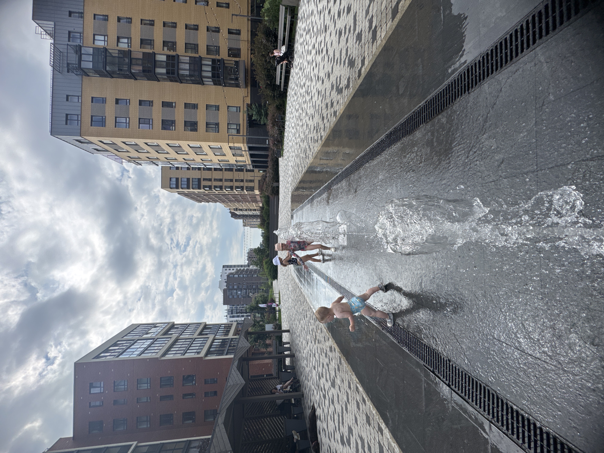 In Novoselye, Russia, a public outdoor space features a low-lying water fountain with children playing in it. In the foreground, a toddler wearing blue patterned swim trunks stands in the shallow water, facing away from the camera and looking towards two older children. Behind the toddler, two girls in swimsuits, one with a white hat and the other with a pink hat, are also interacting with the fountain.
The fountain is situated on a dark, polished surface with visible drainage grates along its edges. The surrounding area is paved with light-colored interlocking stones arranged in a starburst pattern. On the left side of the image, a covered seating area with benches and tables is visible, with a woman and a baby stroller in the background. On the right side, a row of benches faces a yellow-brick apartment building, and a woman is seated on a bench with a stroller.
Beyond the immediate plaza, several multi-story apartment buildings of varying architectural styles are visible, with a cloudy sky overhead. The environment suggests a modern residential complex with recreational amenities.