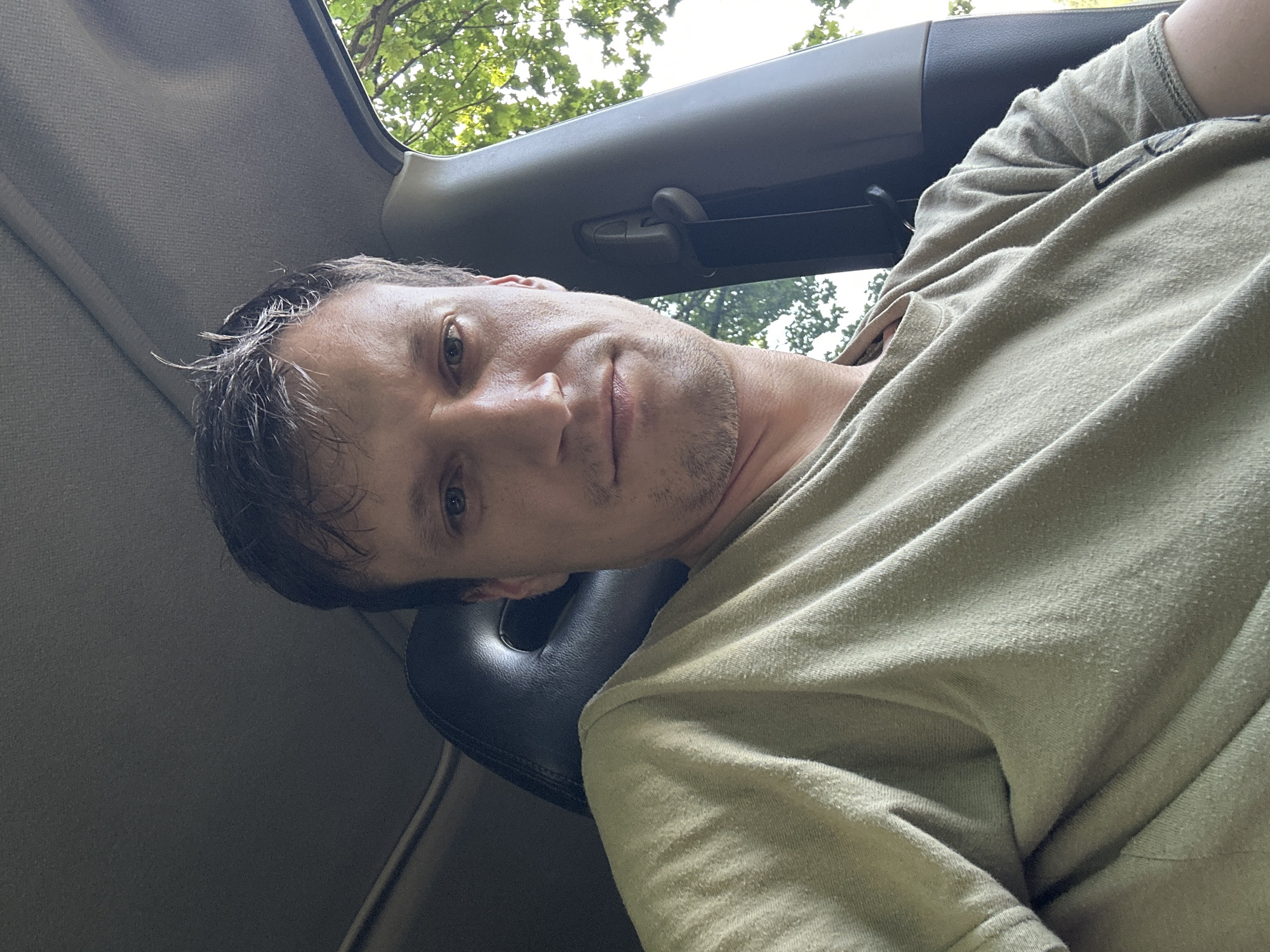 A close-up, low-angle, outdoor selfie taken from the interior of a vehicle shows a man with short, dark, messy hair and blue eyes. He is wearing a light olive-green t-shirt. The man's head and shoulders are visible, with the interior of the car, including the headrest of a dark-colored seat and the car's ceiling, in the foreground. Sunlight filters through trees visible outside the car window on the right side of the frame. The specific location in Makiv, Ukraine, is not discernible from the visual information provided in the image.