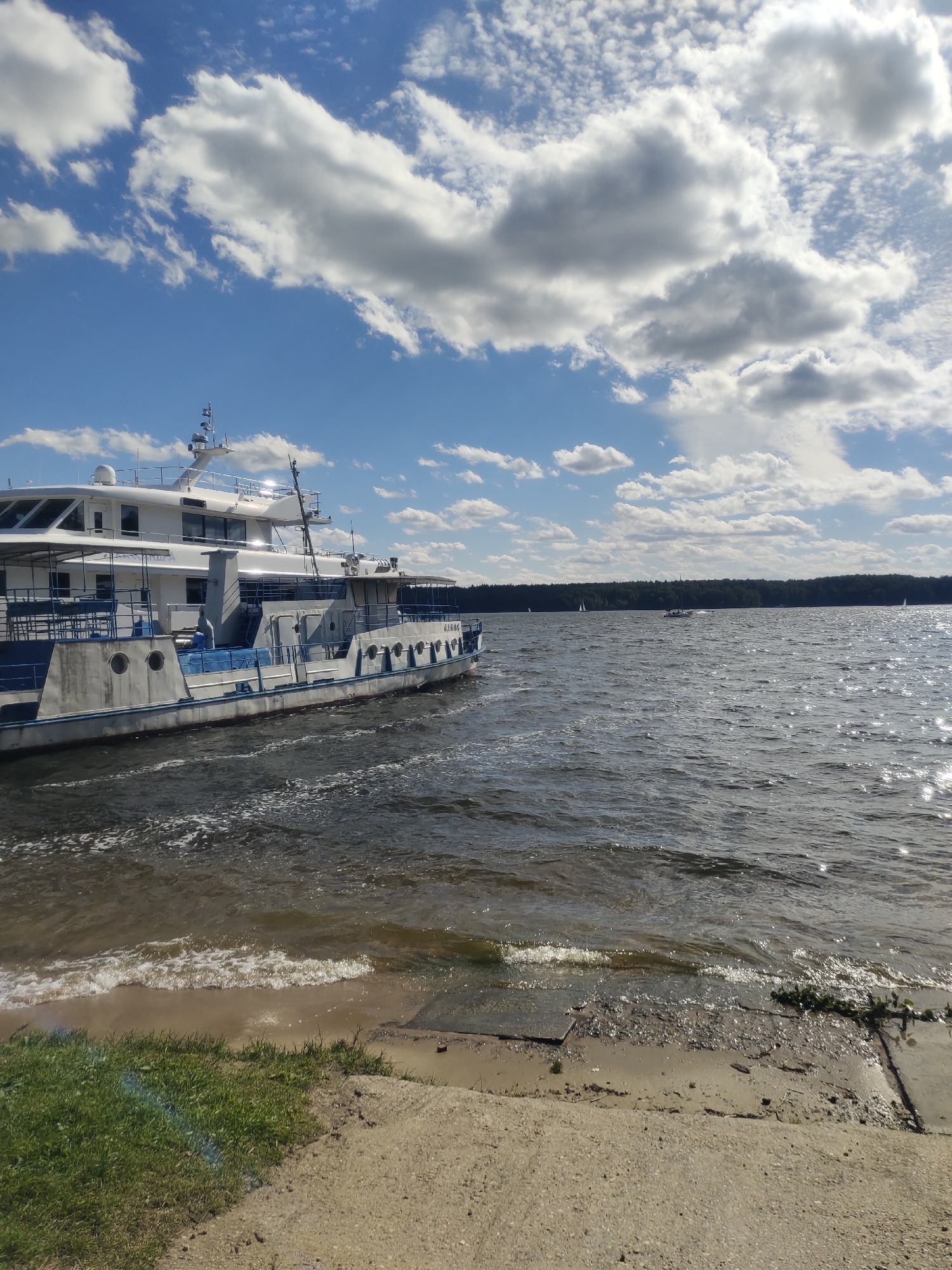 A white and blue passenger boat is docked on the shore of a lake. The boat is a multi-level vessel with a white upper deck and a lower hull. The water is choppy and reflects the sunlight, and there are whitecaps near the shore. The shore is made of sand and concrete, with some grass on the left side of the image. In the distance, across the lake, there is a dense line of green trees. The sky is blue with scattered white clouds. Several small sailboats are visible on the water in the distance. The scene is likely located at Lake Biserovo, in the village of Zhostovo, Russia, known for its lacquer painting art.