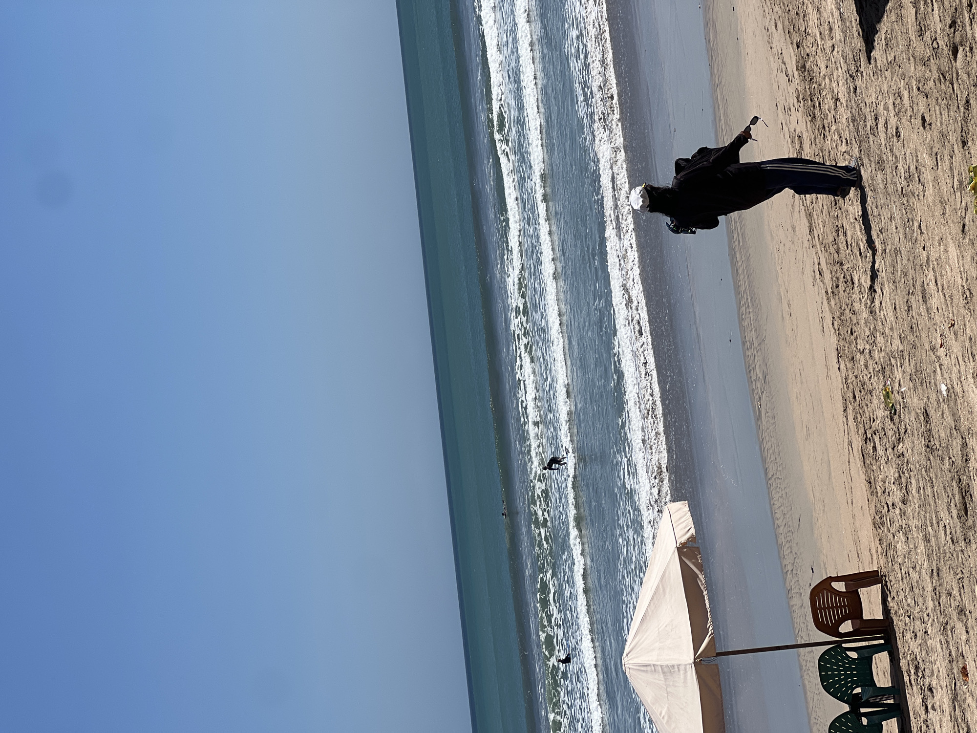 This image depicts a beach scene in Kuta, Indonesia, on a clear, sunny day. The vast expanse of the ocean dominates the background, with several surfers visible in the waves. The water is a deep blue, transitioning to lighter shades near the shore where white-capped waves crash onto the sand. In the foreground, a beach umbrella provides shade over an area with three plastic chairs: two green and one brown. A person wearing a black hooded jacket, dark pants with blue stripes, and a white cap is walking on the sand, holding a camera and sunglasses. The sand is light brown and textured, with some debris visible. The sky is a uniform, bright blue.