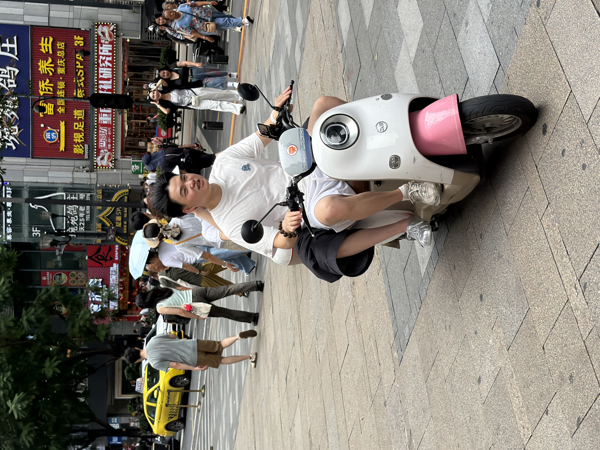 In Yuzhong District, China, a young man wearing a white t-shirt and white shorts rides a white and pink electric scooter on a paved city street. He is wearing white socks and white sneakers and has a bracelet on his left wrist. A person with dark hair, wearing a black skirt and white socks, is seated behind him. The scooter has a white body with a pink fender and a round headlight. The background shows a busy street with buildings, trees, and other pedestrians. A yellow taxi is visible on the left side of the image, and various signs with Chinese characters adorn the buildings. The overall scene depicts a typical urban environment in China.