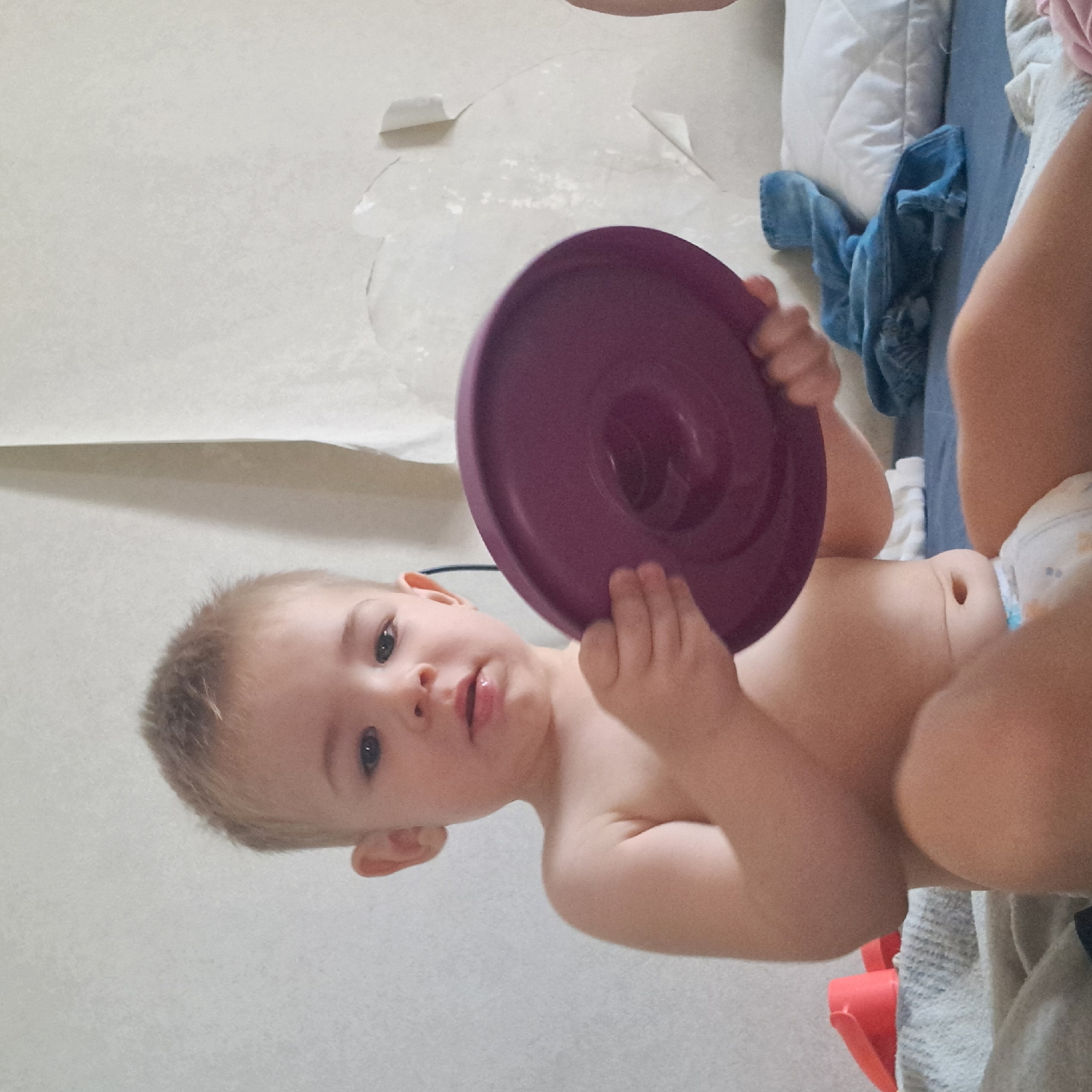 A young child, likely a toddler, is seated on a bed. The child has blonde hair and is looking forward, holding a purple disc-shaped toy in their hands. The child is wearing a diaper. The background consists of a wall with peeling wallpaper and a bed with pillows and a blue blanket. There are also a couple of red plastic toys visible on the left side of the bed. The scene appears to be an indoor domestic setting. There is no information in the image to suggest its location in Krasnoyarsk, Russia.