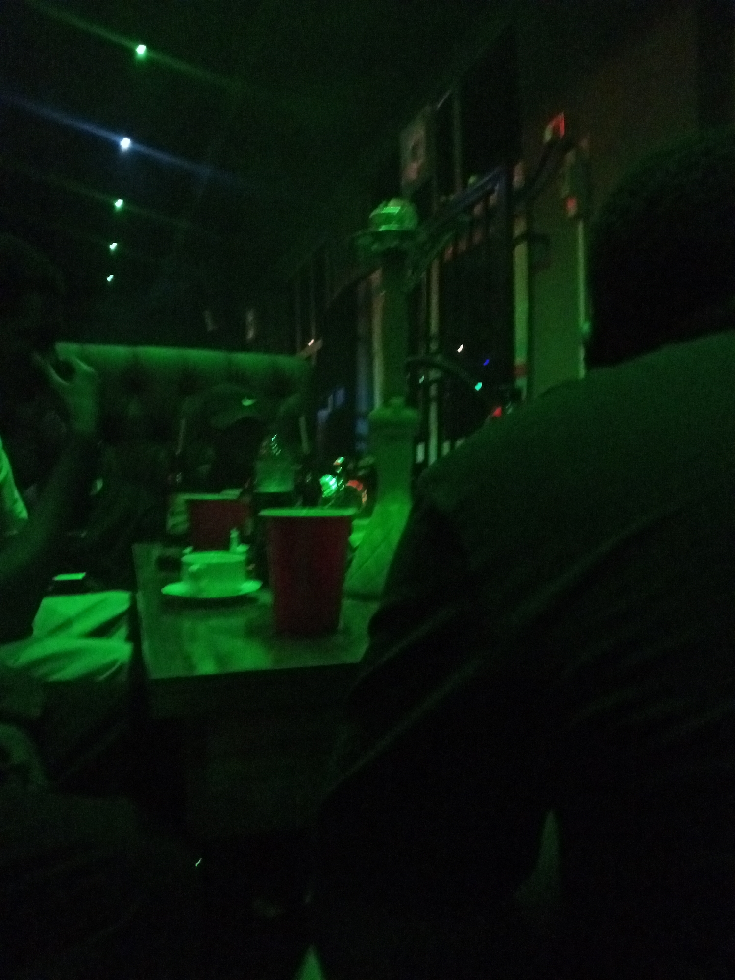The image depicts an indoor scene illuminated by green lighting, suggesting a dimly lit bar or lounge environment, possibly in Lokoja, Nigeria. The focus is on a wooden table with several objects, including red cups and a white cup and saucer. To the left, a person is partially visible, with their arm resting on the table. In the background, other individuals are seated, and the overall ambiance is dark with green light accents coming from overhead fixtures. The scene conveys a casual social gathering, possibly in a recreational establishment.