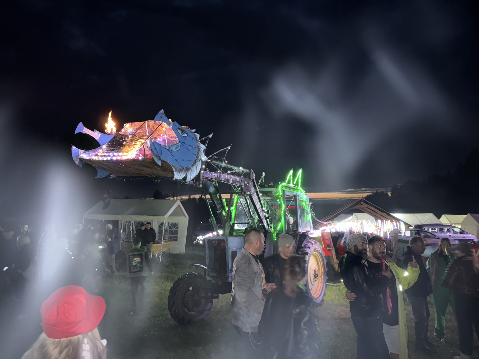This nighttime image depicts an outdoor gathering in Большая Кивара, Russia, centered around a highly customized tractor and various temporary structures. The primary subject is a vintage-style, light blue-grey tractor equipped with a front loader that supports an elaborate, large, illuminated dragon-like head structure. This head is intricately designed with blue, pink, orange, and purple hues, and features an open maw from which a plume of fire is actively erupting upwards. The tractor itself is adorned with bright green LED strip lights along its chassis, cabin, and front loader mechanism.
Numerous individuals are scattered across the grassy foreground and midground, observing and interacting. In the immediate foreground, the back of a person wearing a bright red hat and light-colored hair is visible, facing away from the viewer. Around the tractor, people are standing in small groups, conversing. One man in the midground wears a dark jacket with a white skull design on the back. Another man near the right tire of the tractor wears a bright yellow jacket. The attire of the crowd is predominantly casual, consisting of jackets, t-shirts, and hats.
In the background, several white marquee-style tents are erected, some appearing to be open stalls with items displayed on tables. A light-colored car or van is partially visible behind one of these tents on the right side. The ground beneath the attendees is unpaved, consistent with a field or open outdoor space. The scene is enveloped in plumes of white smoke or fog, illuminated by the artificial lights from the tractor and tents, creating a dynamic and atmospheric visual. The sky above is dark, indicating a clear night.