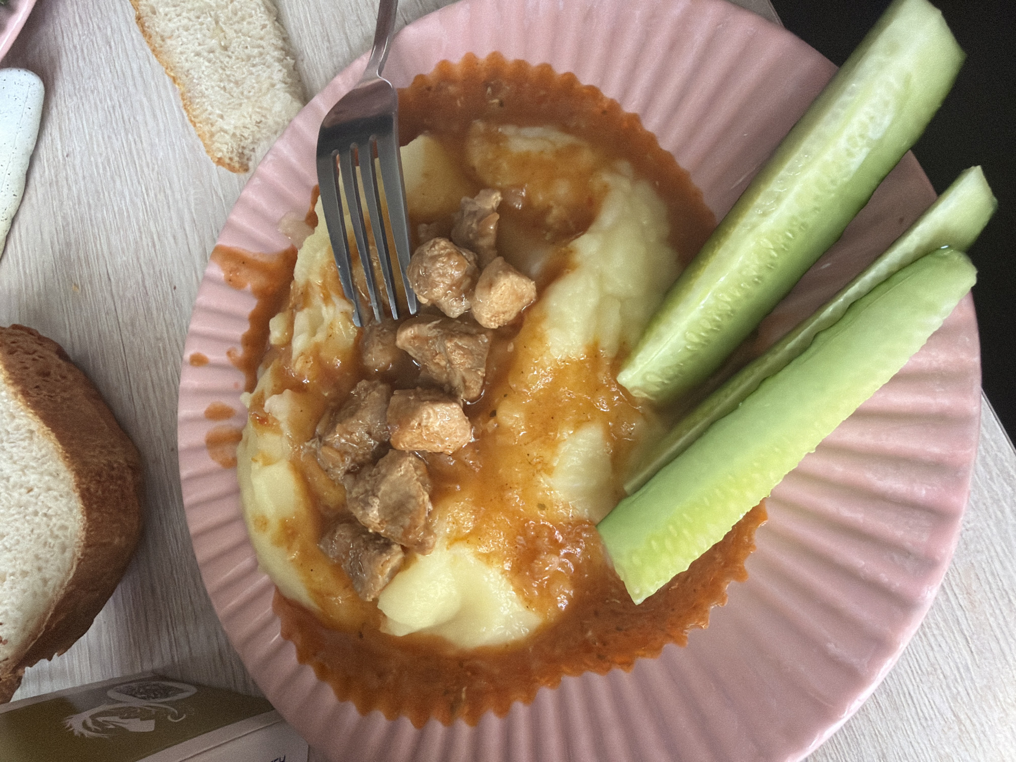 A close-up, overhead shot captures a meal on a pink, fluted plate. The plate contains a serving of mashed potatoes topped with stewed meat in a reddish-orange sauce. Three cucumber sticks are arranged in front of the mashed potatoes. A fork is partially submerged in the mashed potatoes and meat. In the background, two slices of bread and a portion of a carton with a horse logo are visible on a light-colored wooden surface. This scene is not located in the city of Shumki, Russia.