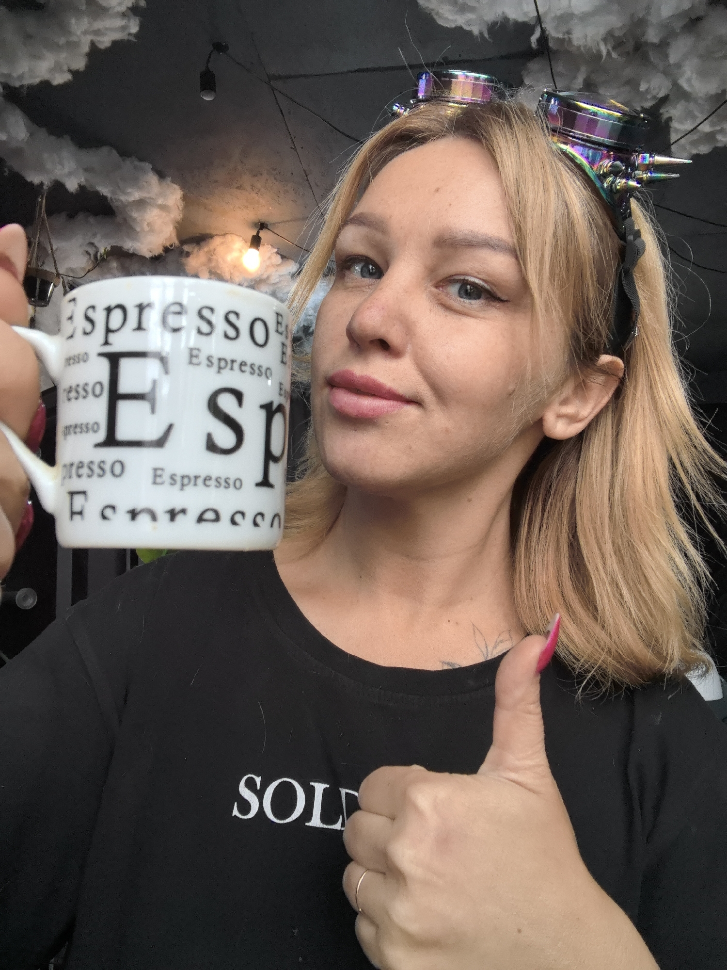 A woman with light blond hair and blue eyes is holding a white mug with the word "espresso" printed on it. She is wearing a black T-shirt with the word "SOLD" printed on it in white letters, and goggles on her head. She is giving a thumbs-up. The background appears to be decorated with artificial clouds. The location is likely not in Noviy, Russia.