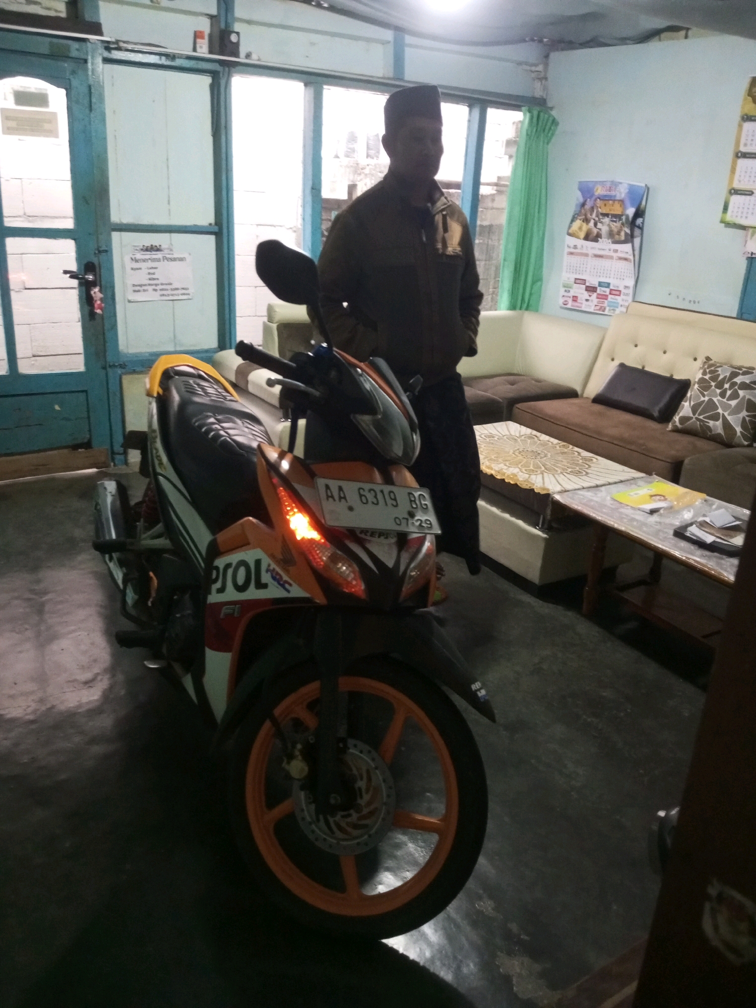 A man stands next to a parked motorcycle inside what appears to be a garage or workshop in Andongsili, Indonesia. The man, wearing a dark jacket and a cap, is positioned to the right of the motorcycle. The motorcycle, predominantly orange and black, is parked facing the viewer. The environment includes a tiled floor, light blue walls, and a white couch in the background. A calendar is visible on the wall, along with a sign on the blue door that reads "Menerima Pesanan" (Accepting Orders). The lighting is dim, suggesting an indoor setting.